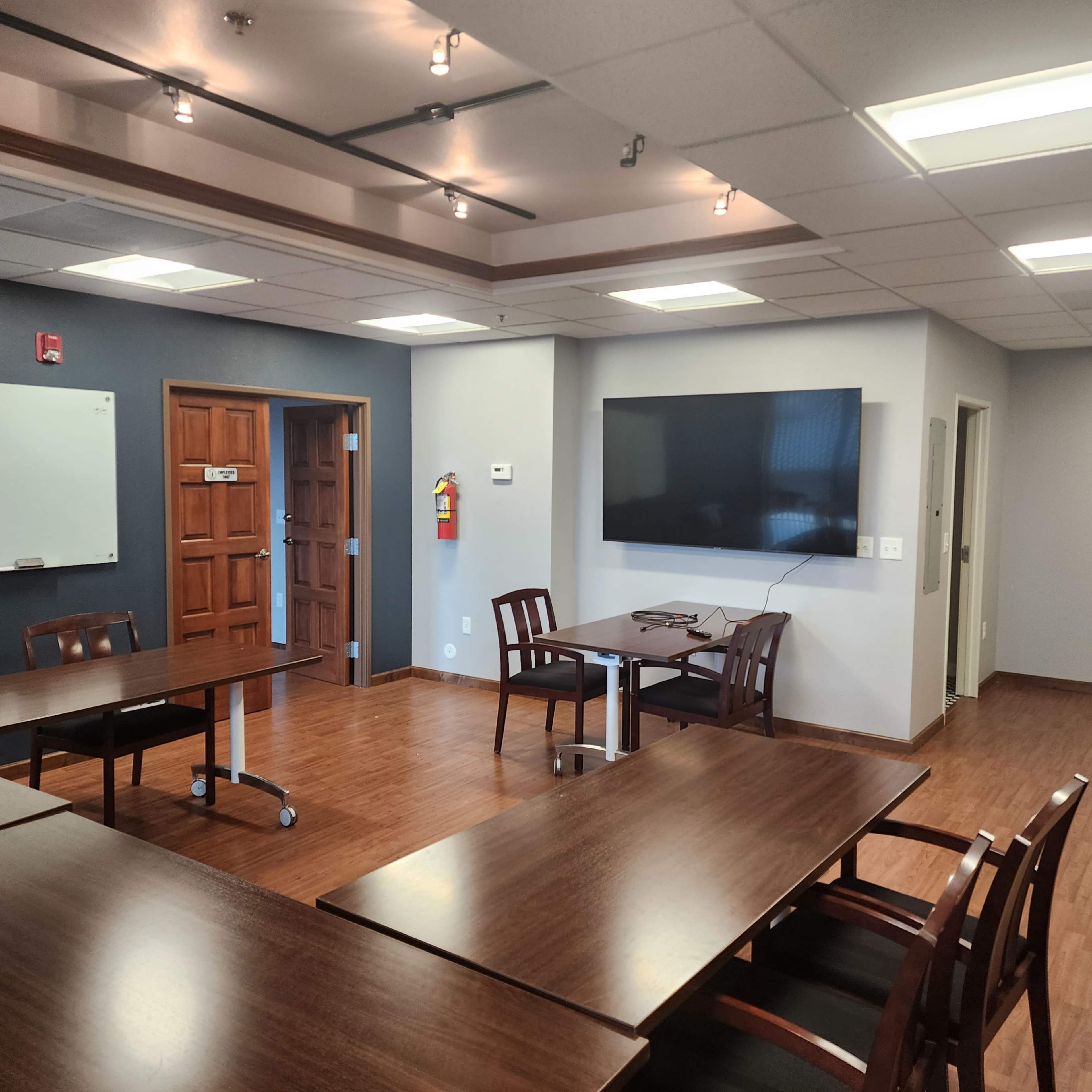A meeting room with several wooden tables and chairs, a whiteboard, and a wall-mounted television.