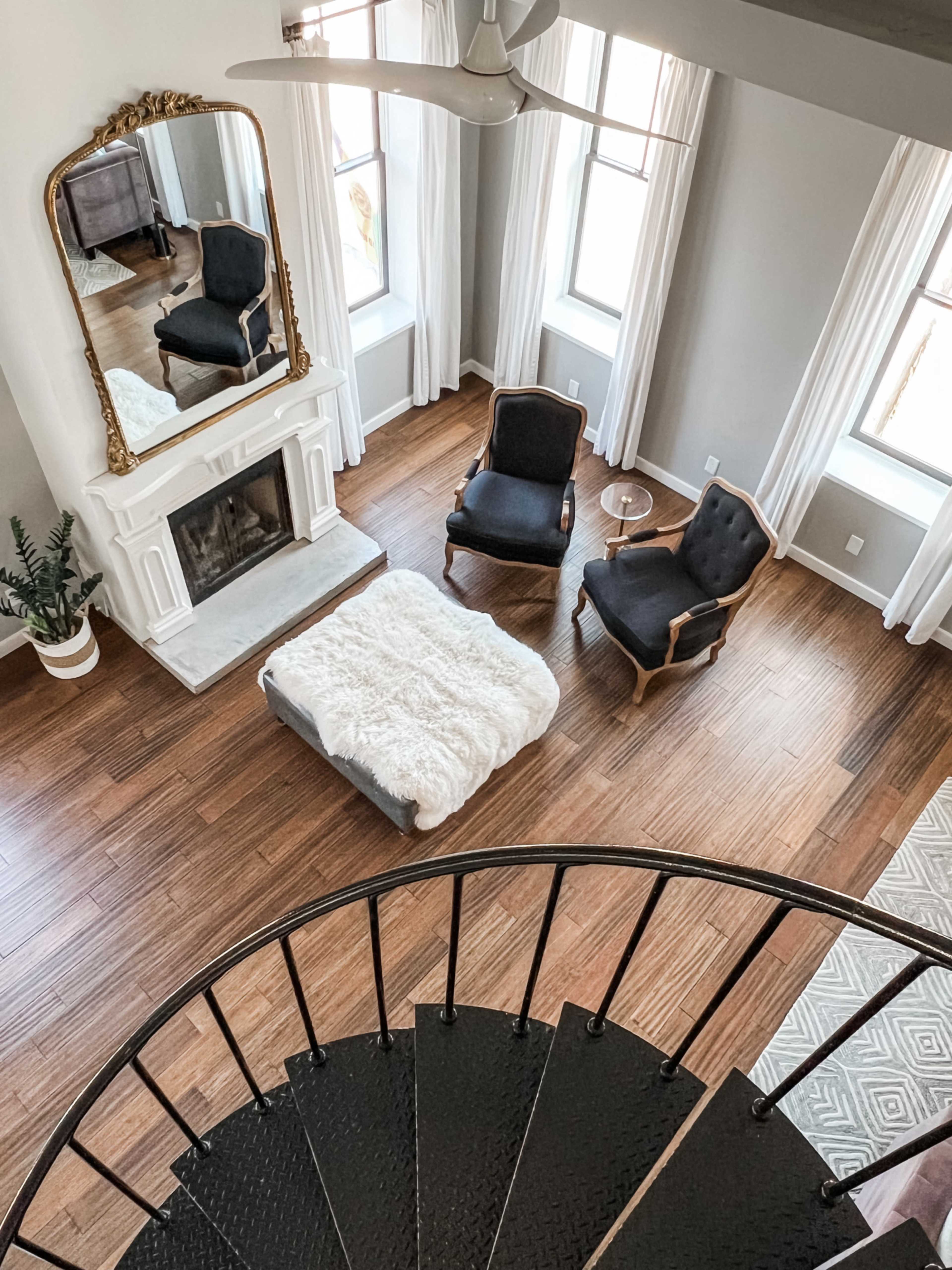 The image shows a modern living room with a white fireplace, two black armchairs, a circular coffee table covered with a throw blanket, and large windows adjacent to a spiral staircase.