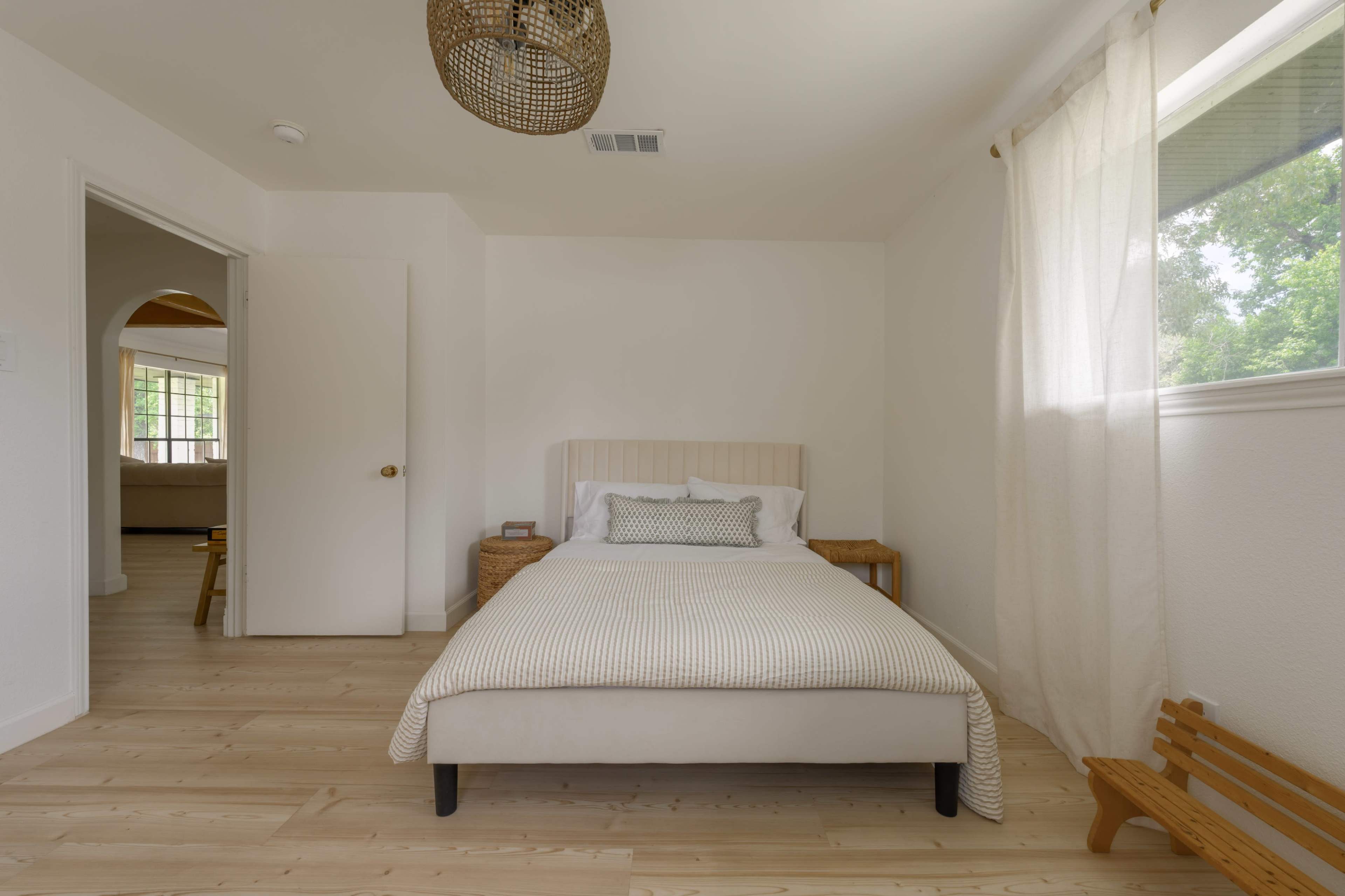 The image shows a simple, well-lit bedroom featuring a bed with a patterned cover, a woven basket, and a small wooden bench.