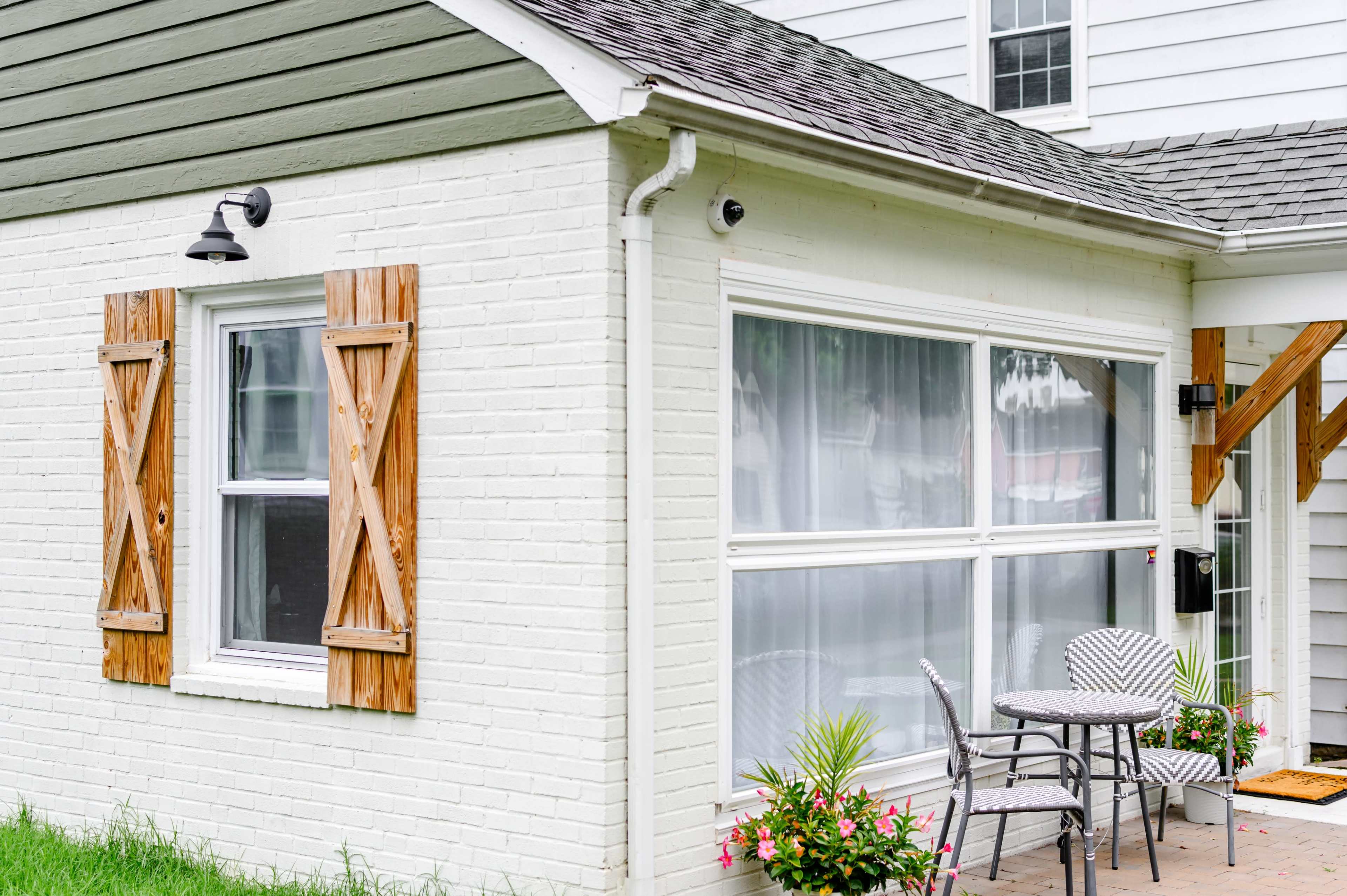The image features a house exterior with white brick walls, wooden shutters, a small table and chairs on a patio, and potted plants.