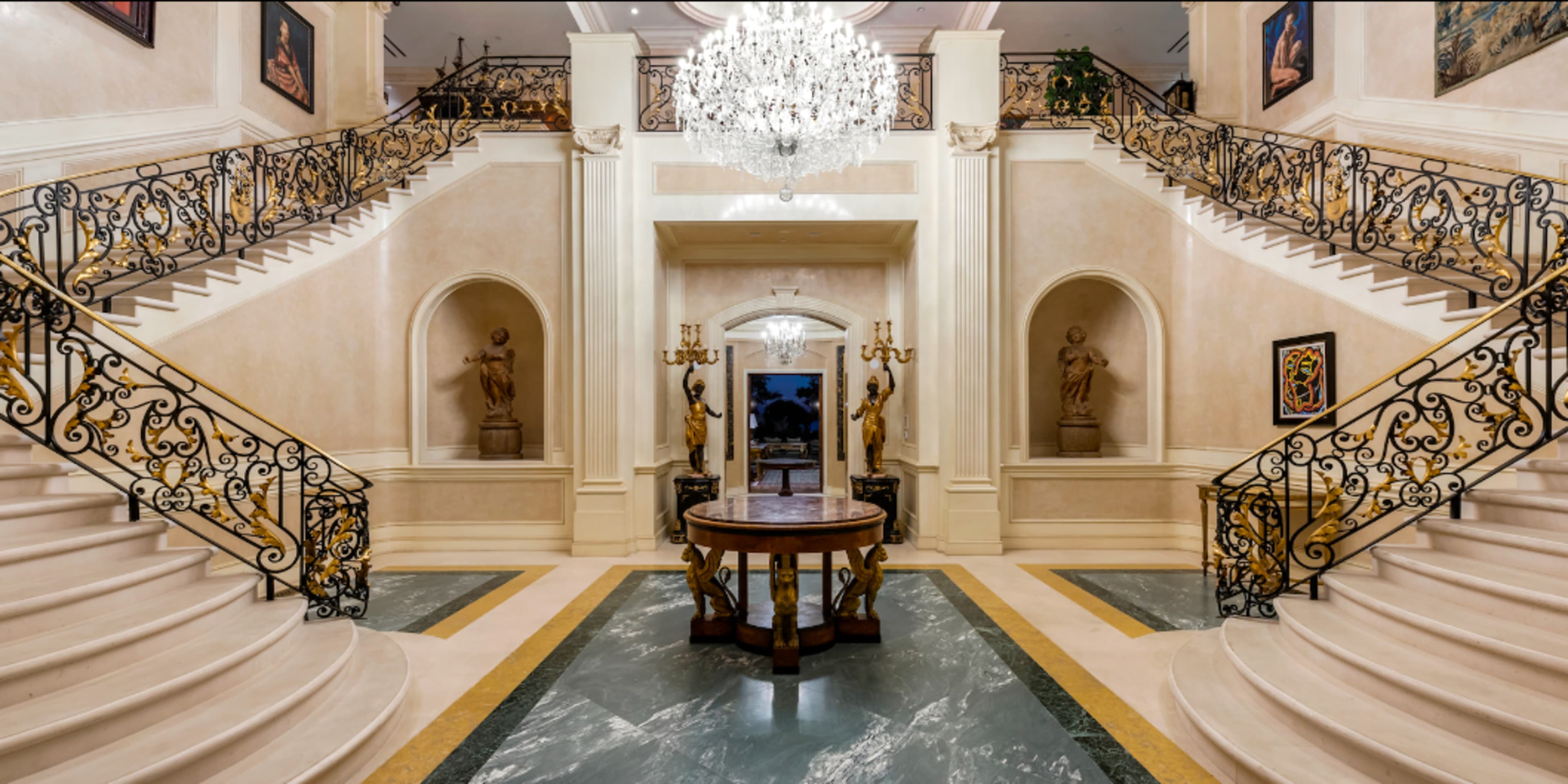 The image shows an ornate foyer with a large chandelier, a round table in the center, and grand staircases on either side, each adorned with decorative railings and statues.