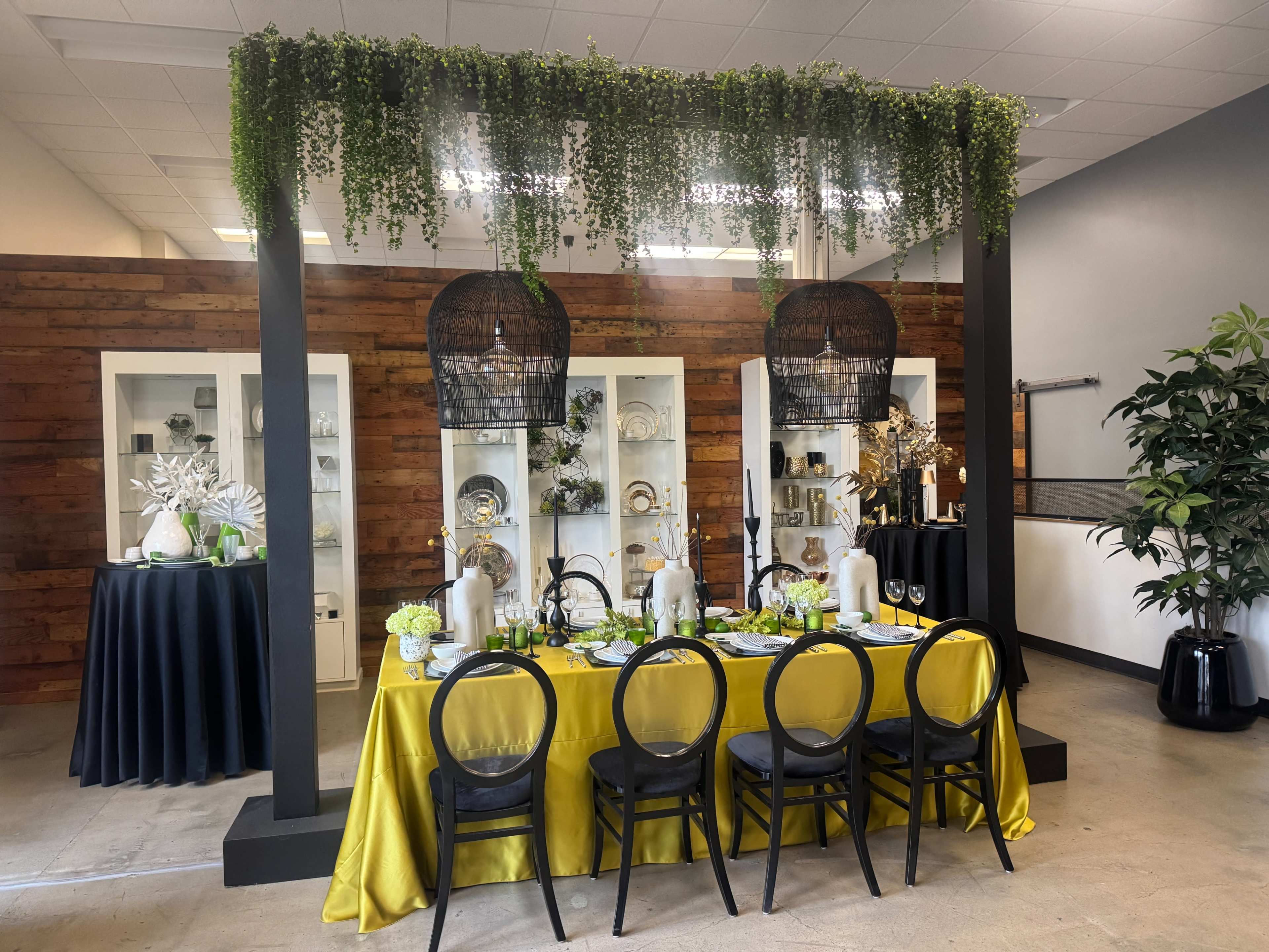 A dining setup features a vibrant yellow tablecloth with black chairs, surrounded by decorative glassware and plants, under a canopy of greenery.