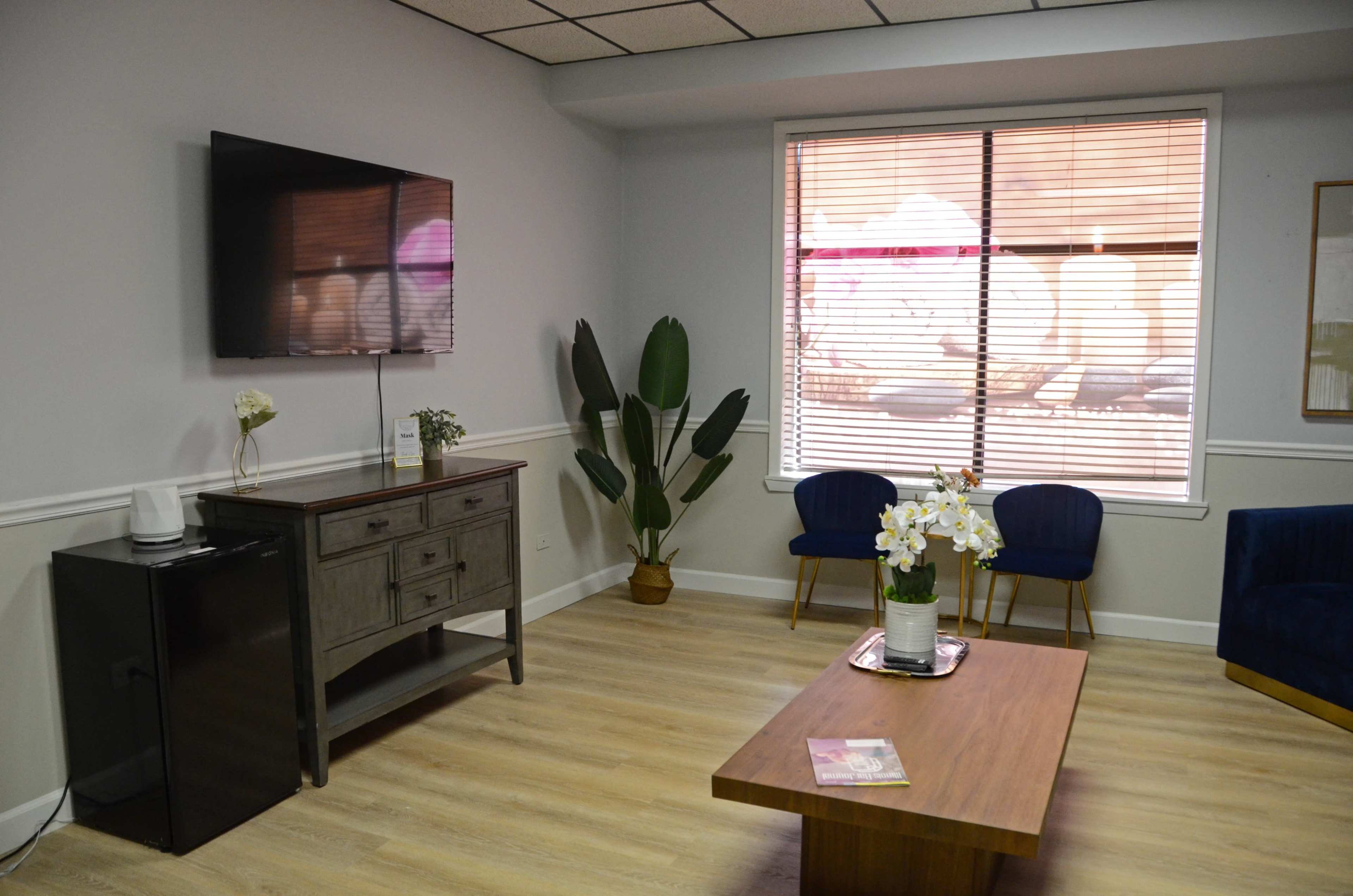 A modern waiting room with a wall-mounted TV, a wooden coffee table, a plant, and two blue upholstered chairs near a window with closed blinds.