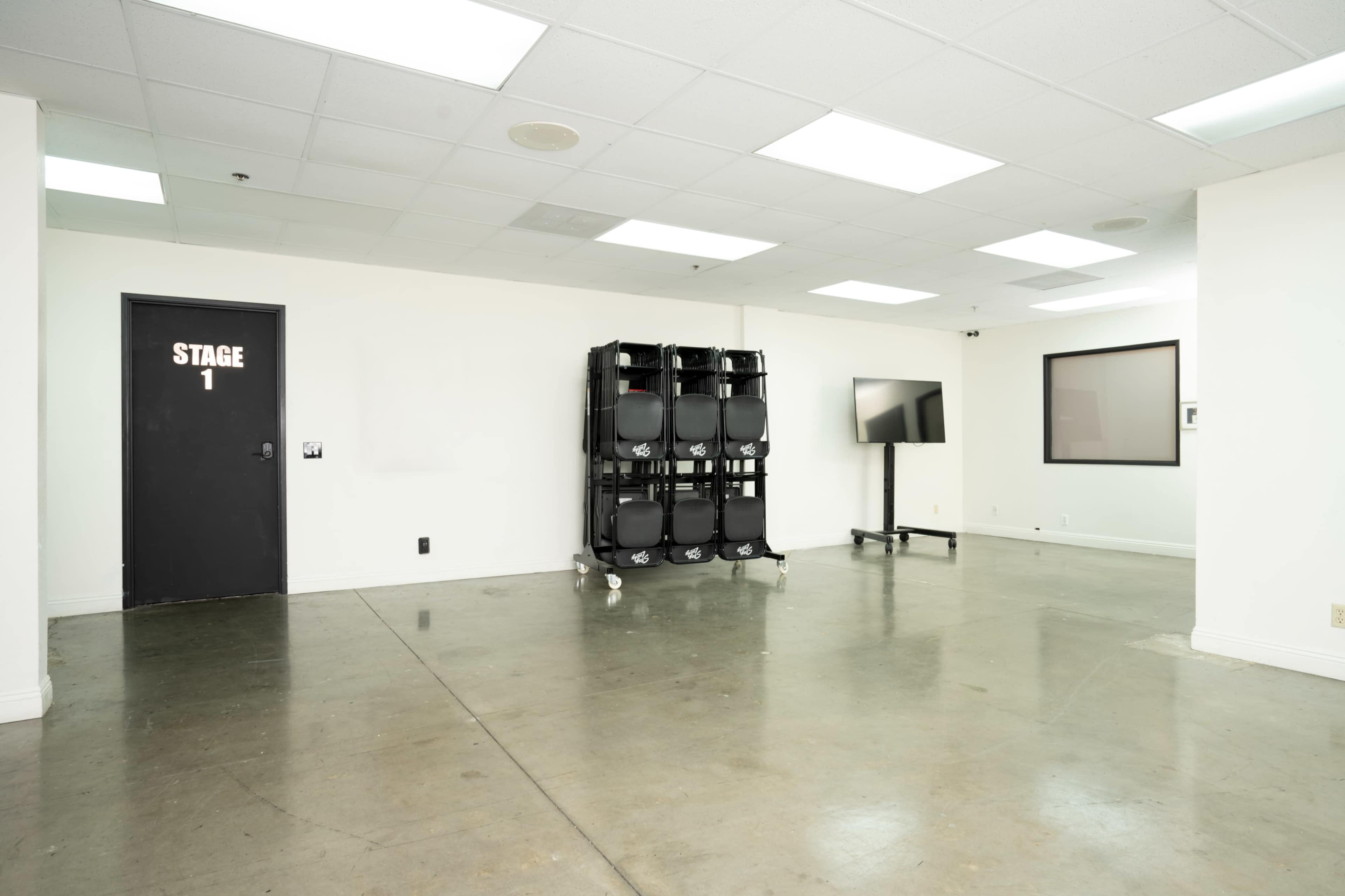 The image shows a minimalist room with a set of stacked black chairs, a television on a stand, and a door labeled "STAGE 1."