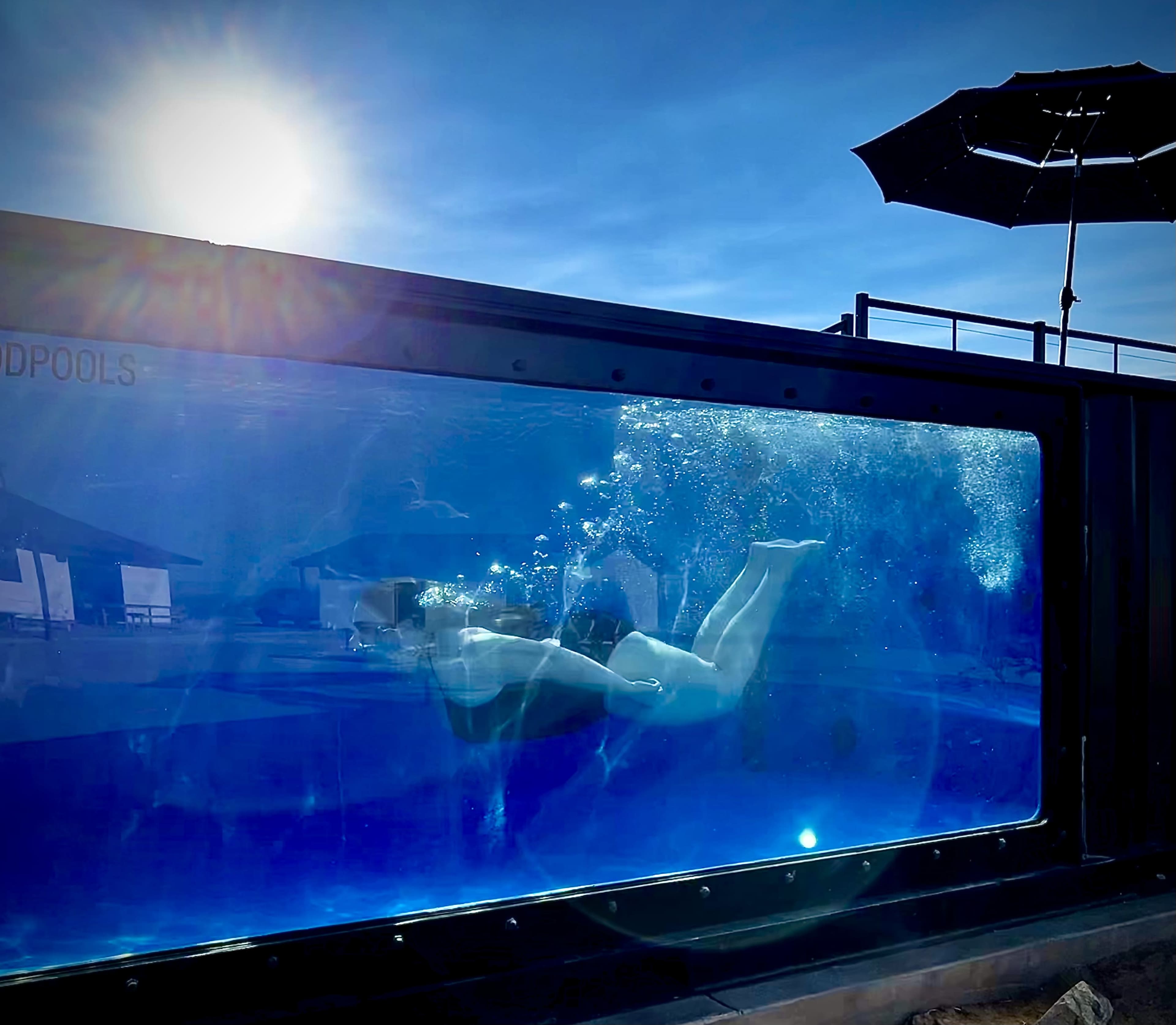 Two swimmers are underwater inside a large glass pool with sunlight shining above and an umbrella in the background.