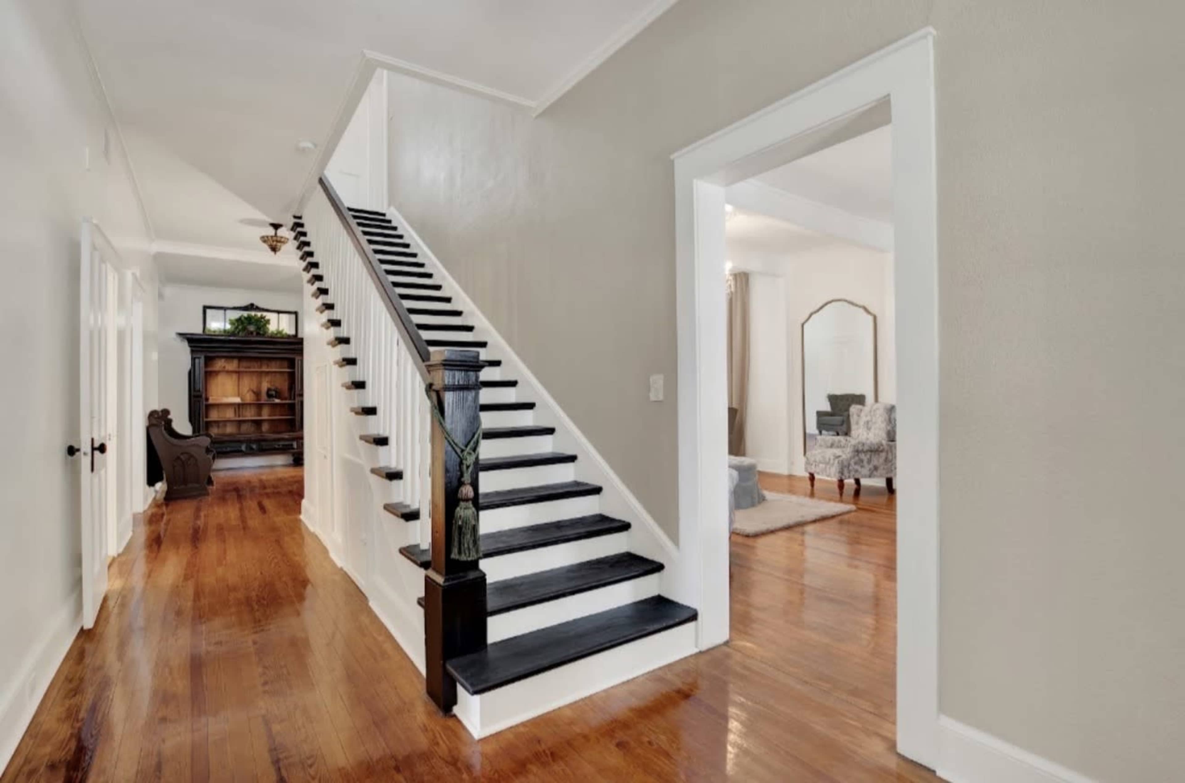 The image shows a hallway featuring a staircase with a black and white design, hardwood floors, and an open doorway leading to a separate room.