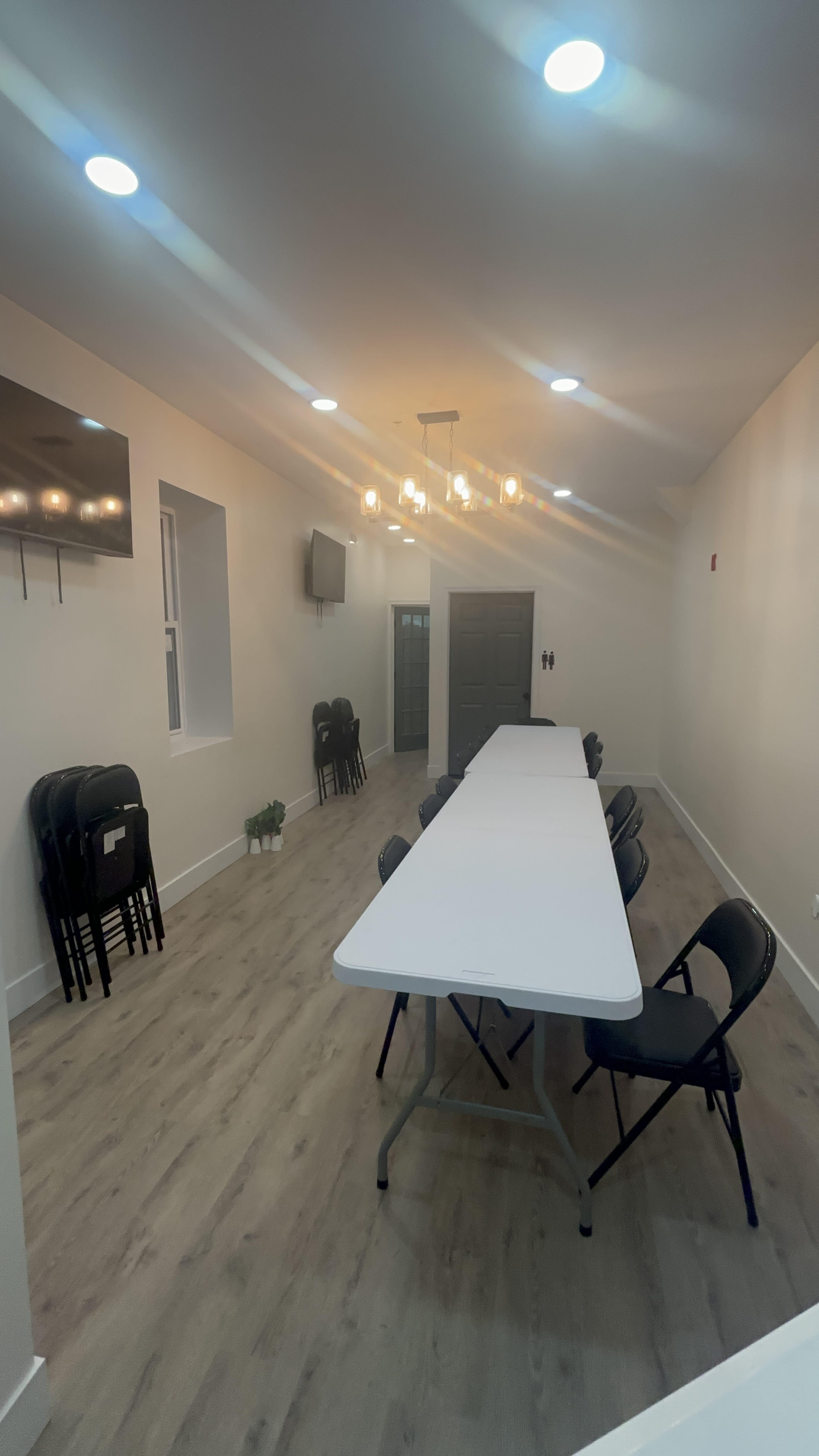 The image shows a spacious meeting room with a long rectangular table, black chairs, wall-mounted TVs, and a light fixture with multiple bulbs.