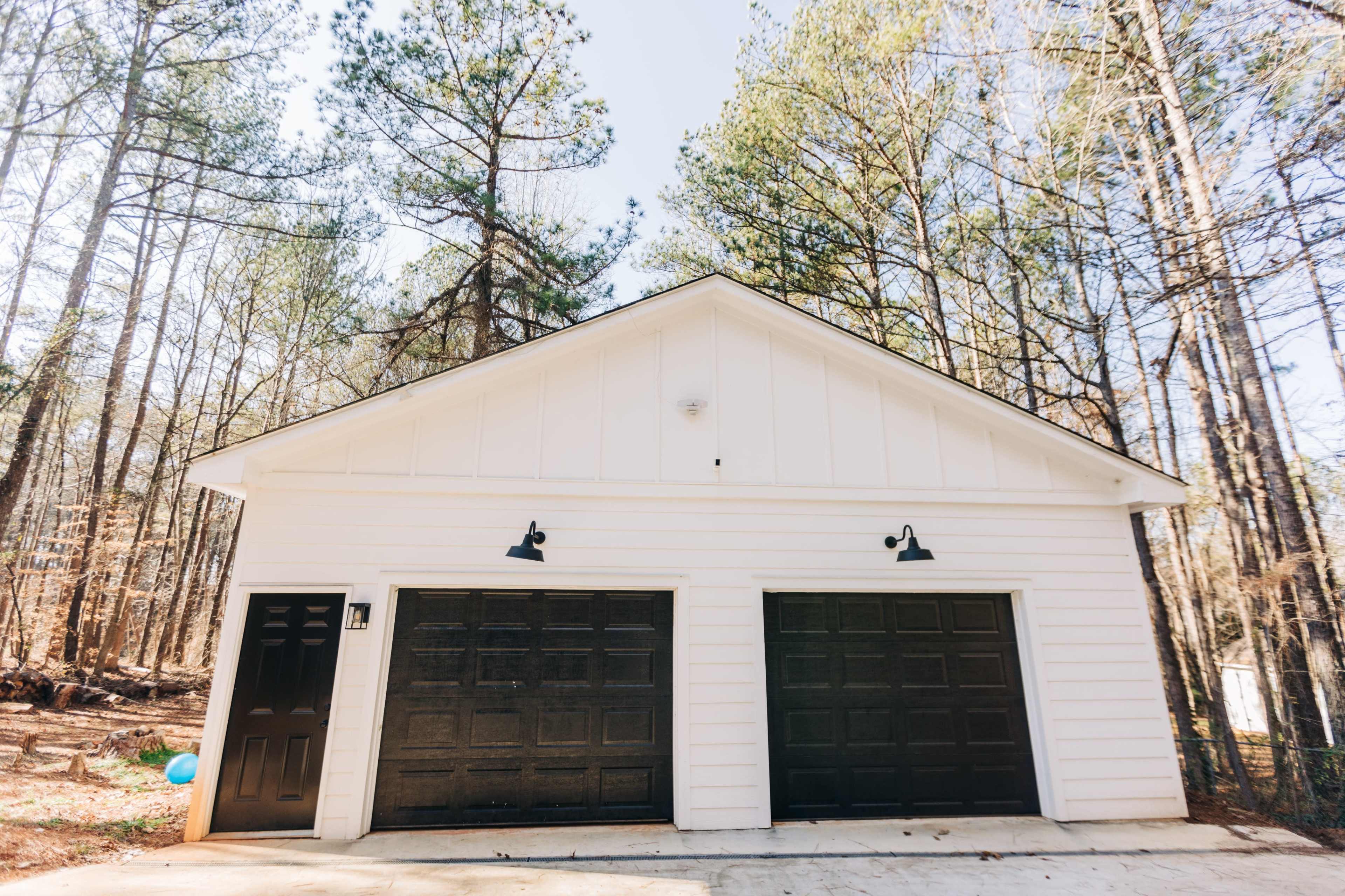 A white garage with two black doors, situated in a wooded area with tall trees in the background.