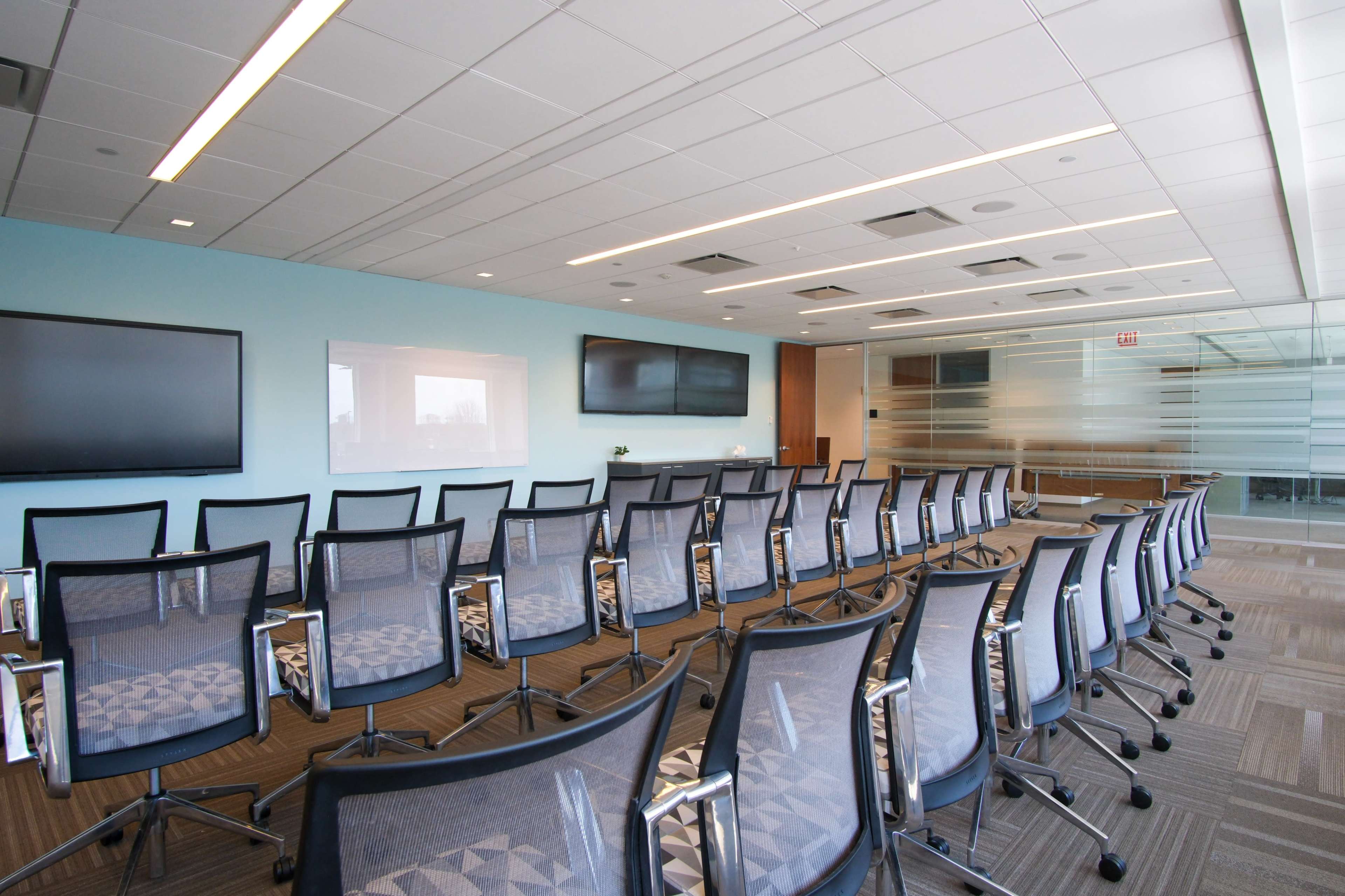 The image shows a modern conference room with rows of chairs facing two large screens and a glass partition.