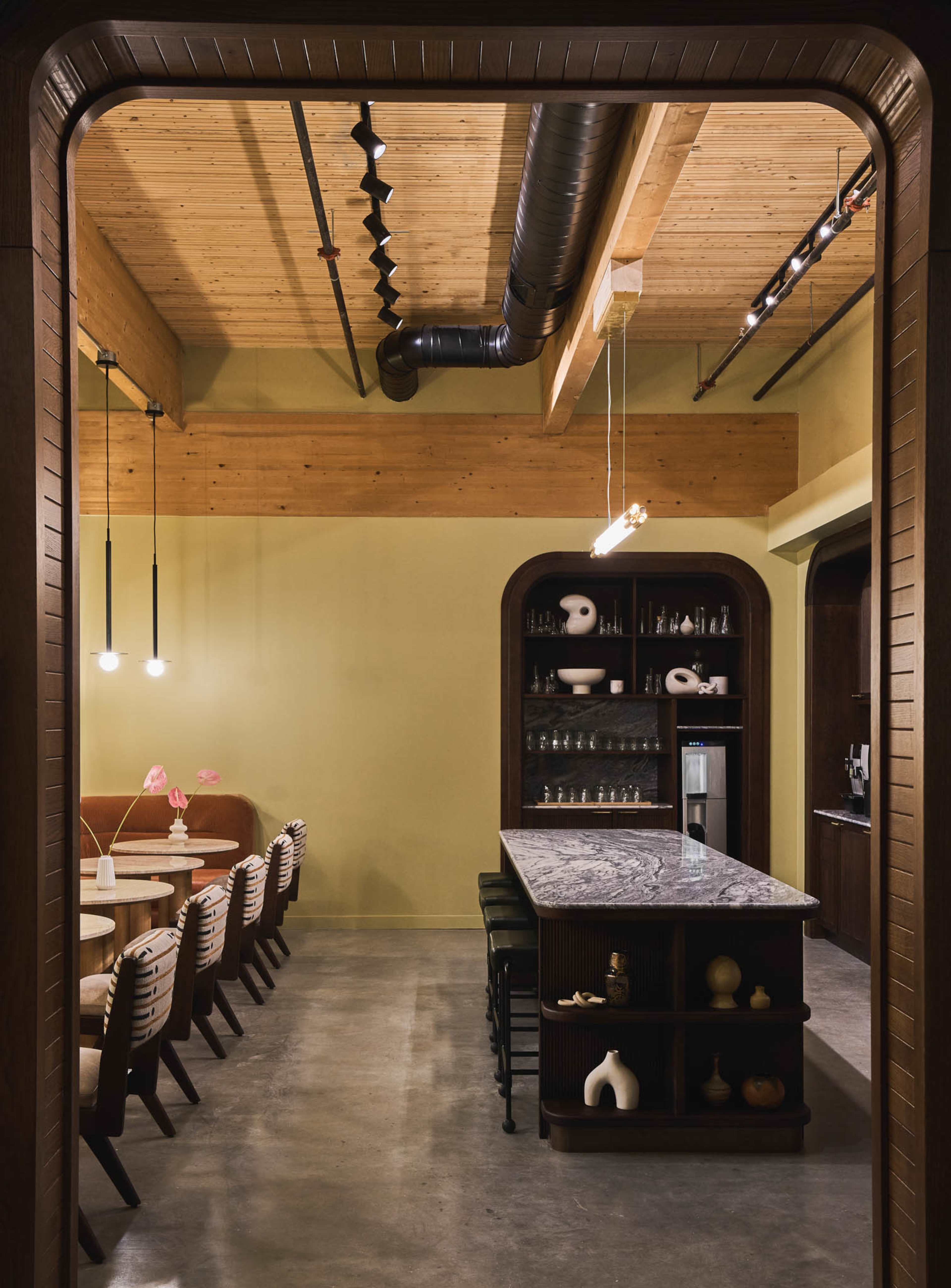 A modern interior space with a long bar counter, seating area, and wooden beams on the ceiling, illuminated by pendant lights.