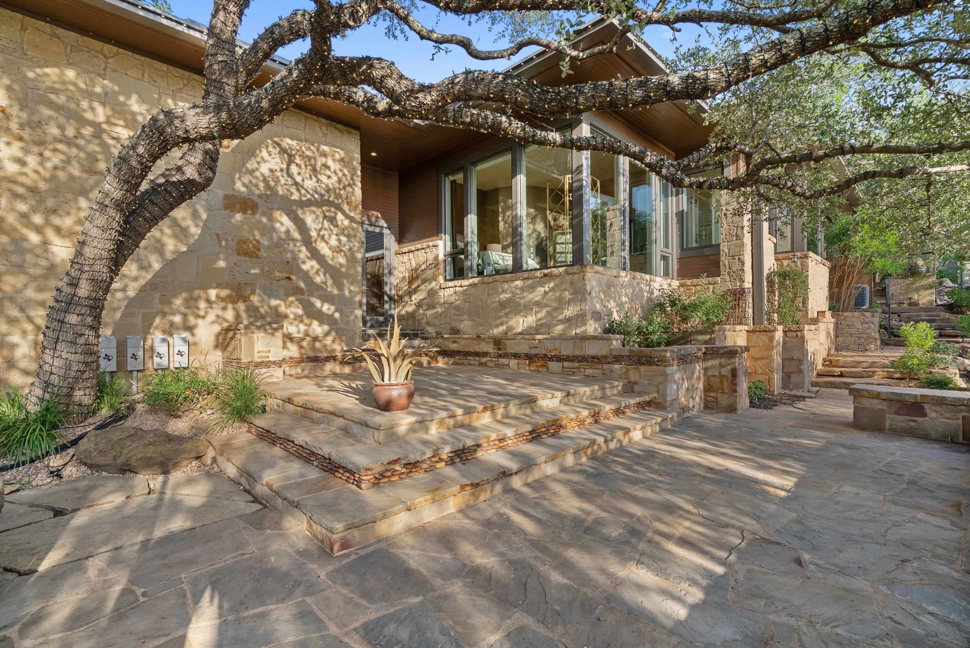 The image shows a house with a stone exterior, featuring large windows and a staircase leading to a landscaped entrance surrounded by trees and plants.