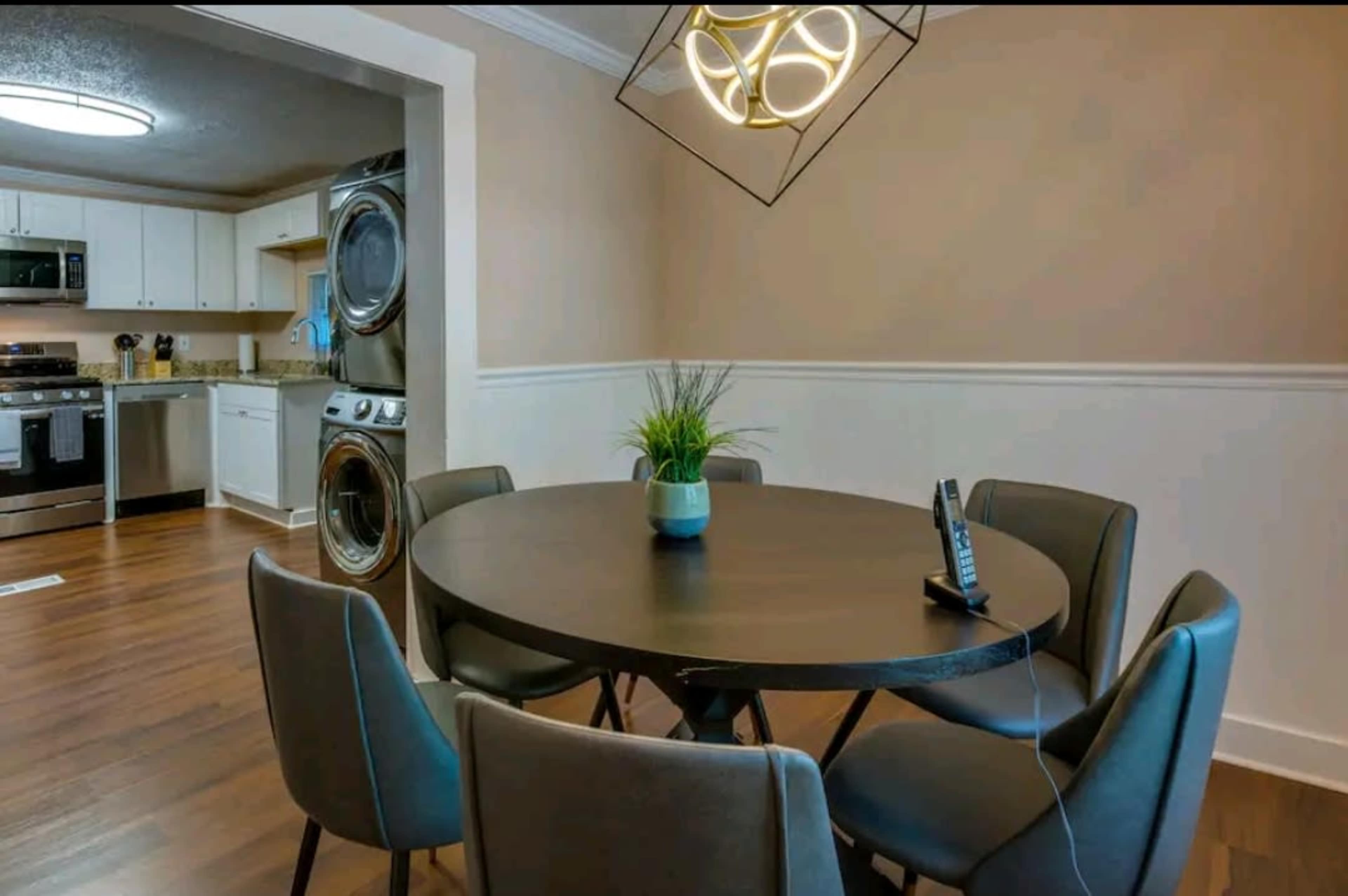 A round dining table with six chairs is set in a kitchen area featuring a washing machine and dryer in the background.