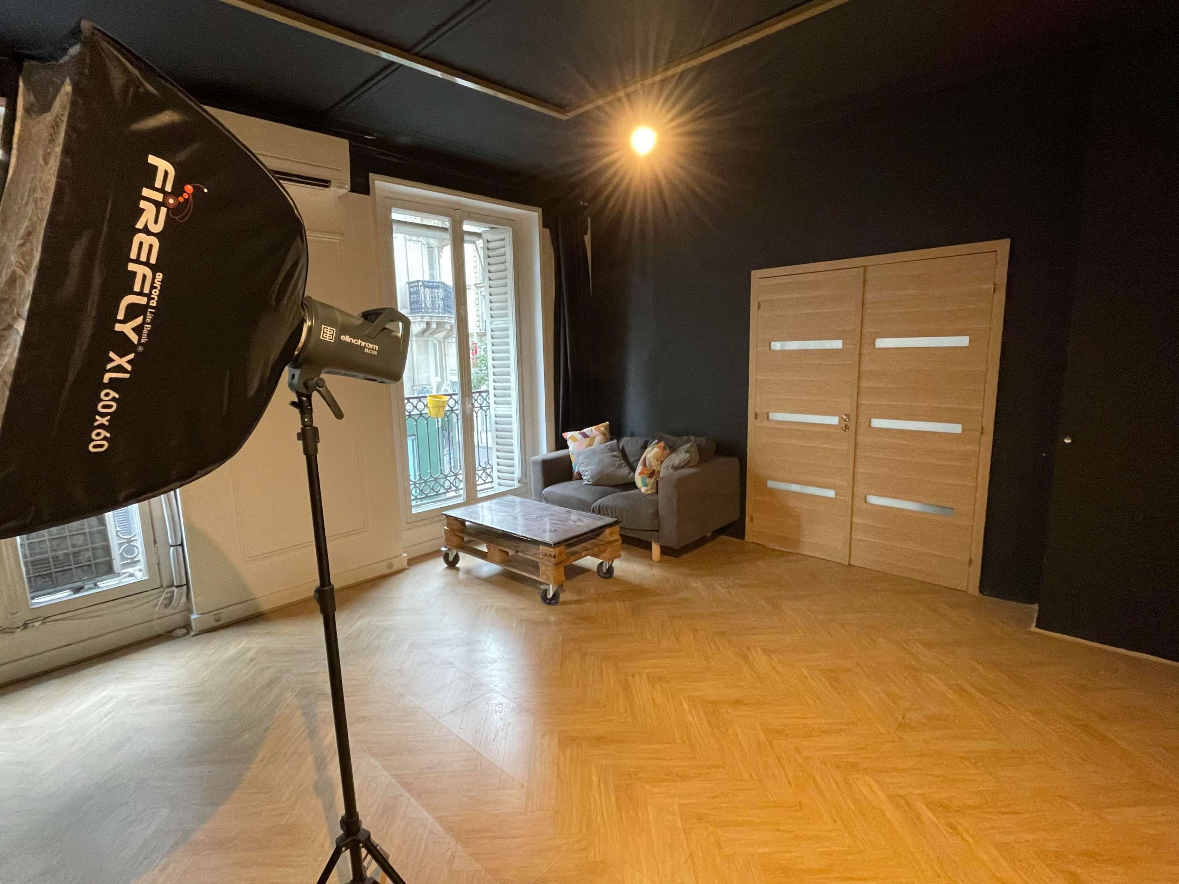 A spacious indoor area with a wooden floor, a coffee table on wheels, a gray sofa, and a large softbox lighting setup directed towards the seating area.