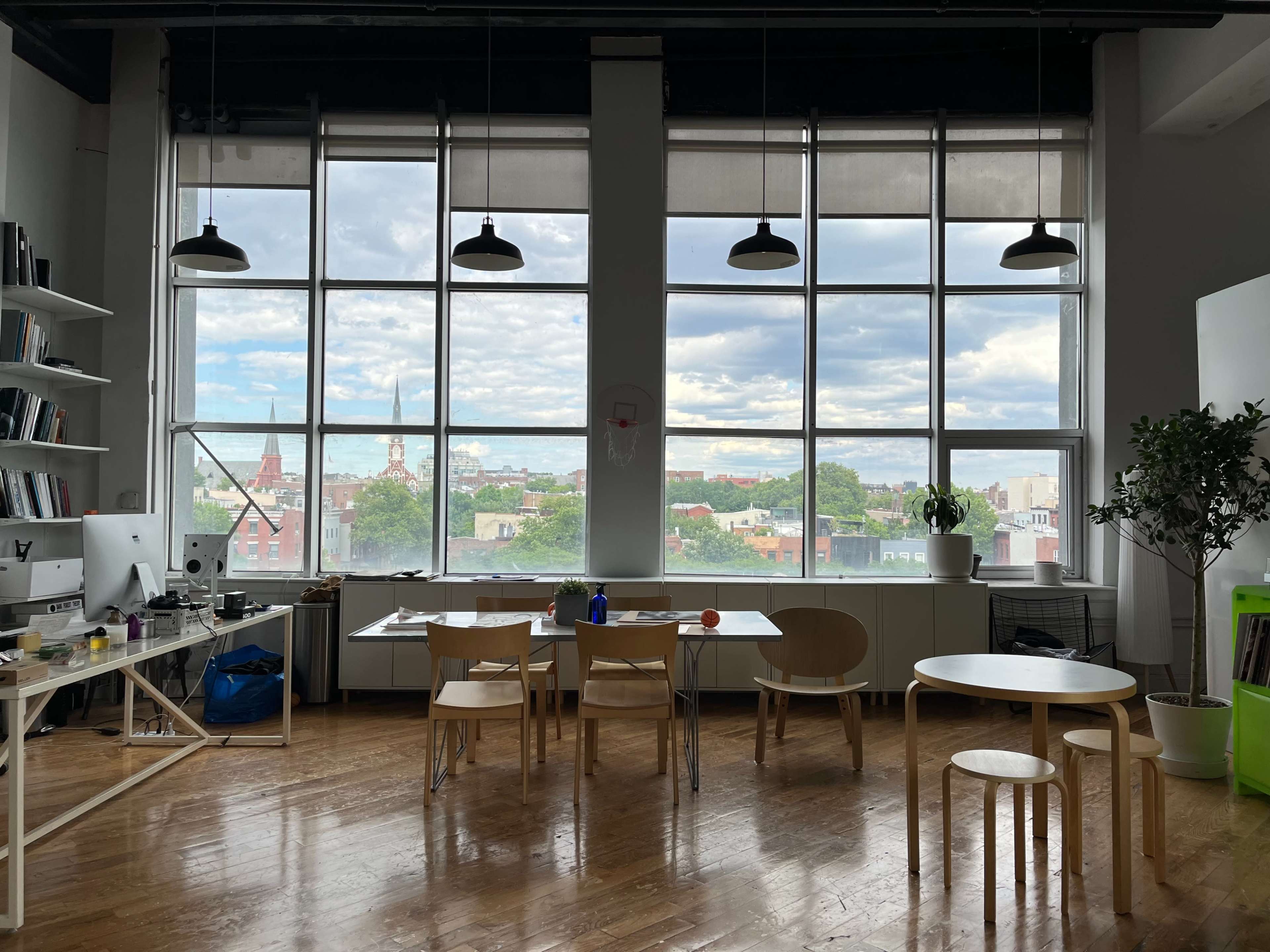 A spacious office features a large window with a view of city rooftops, a wooden table surrounded by chairs, and a bookshelf along one wall.