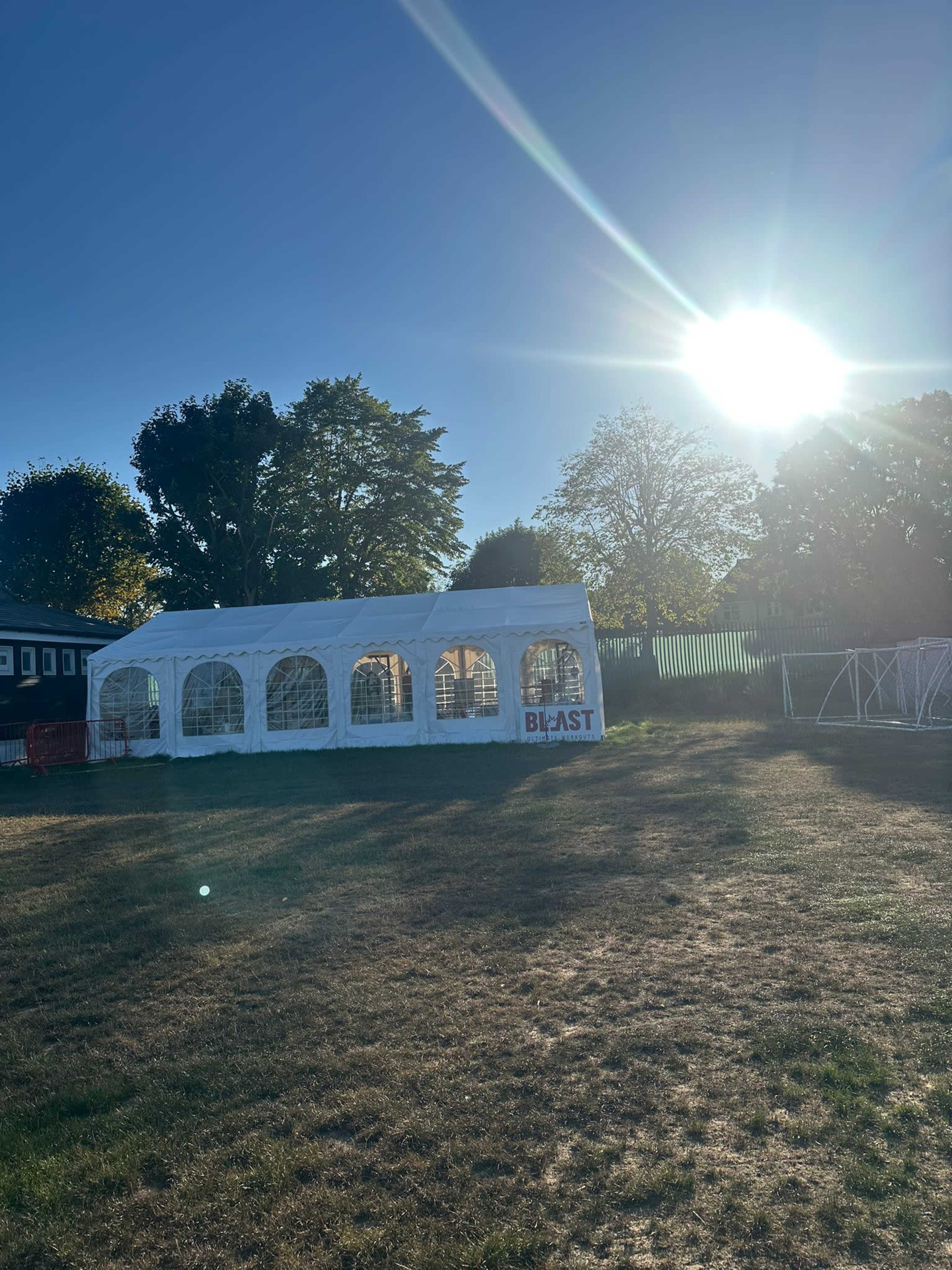 A white tent with large windows stands in an open field under a bright sun, surrounded by trees.