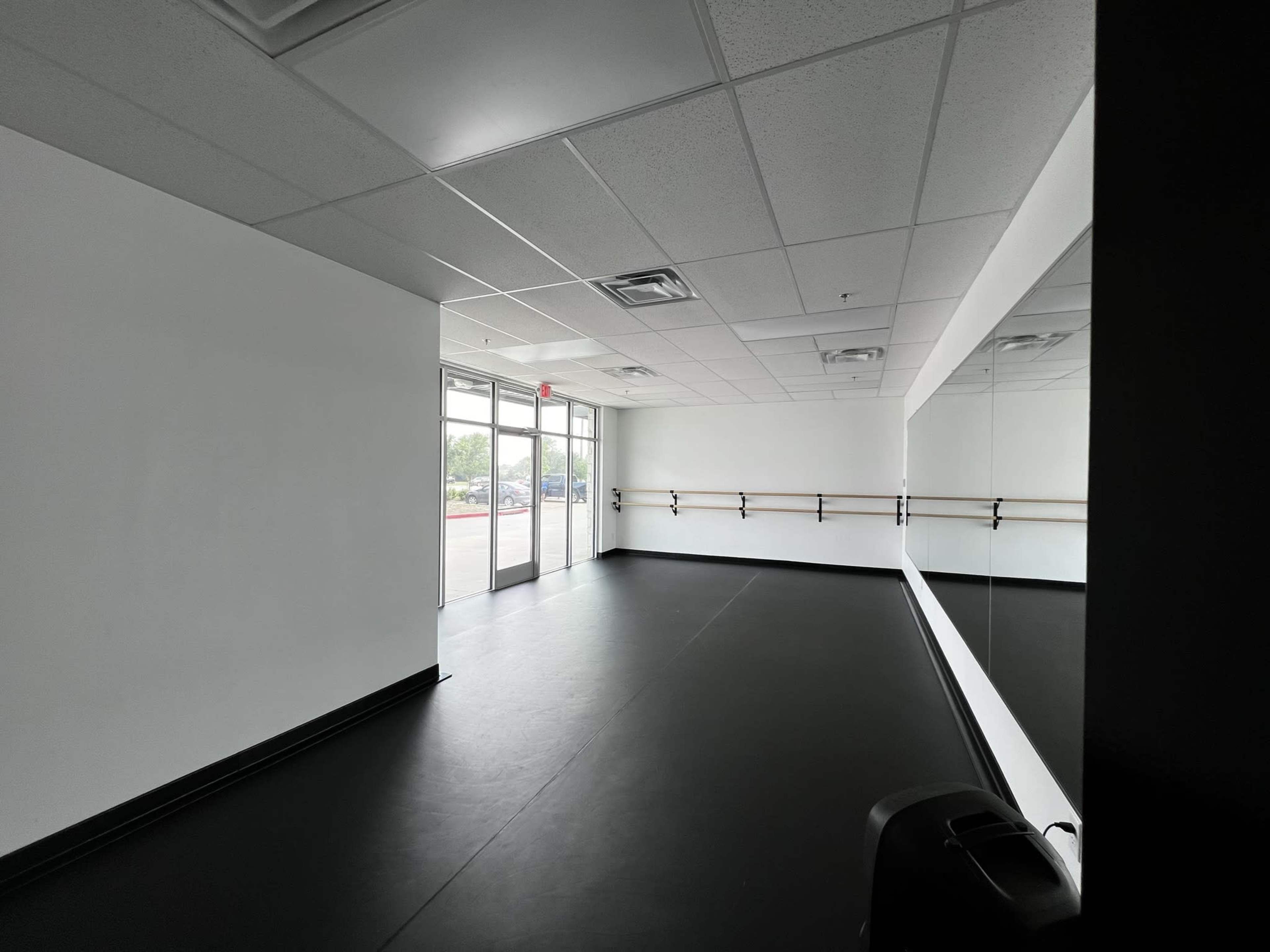 An empty dance studio with black flooring, white walls, and a large mirror along one side.
