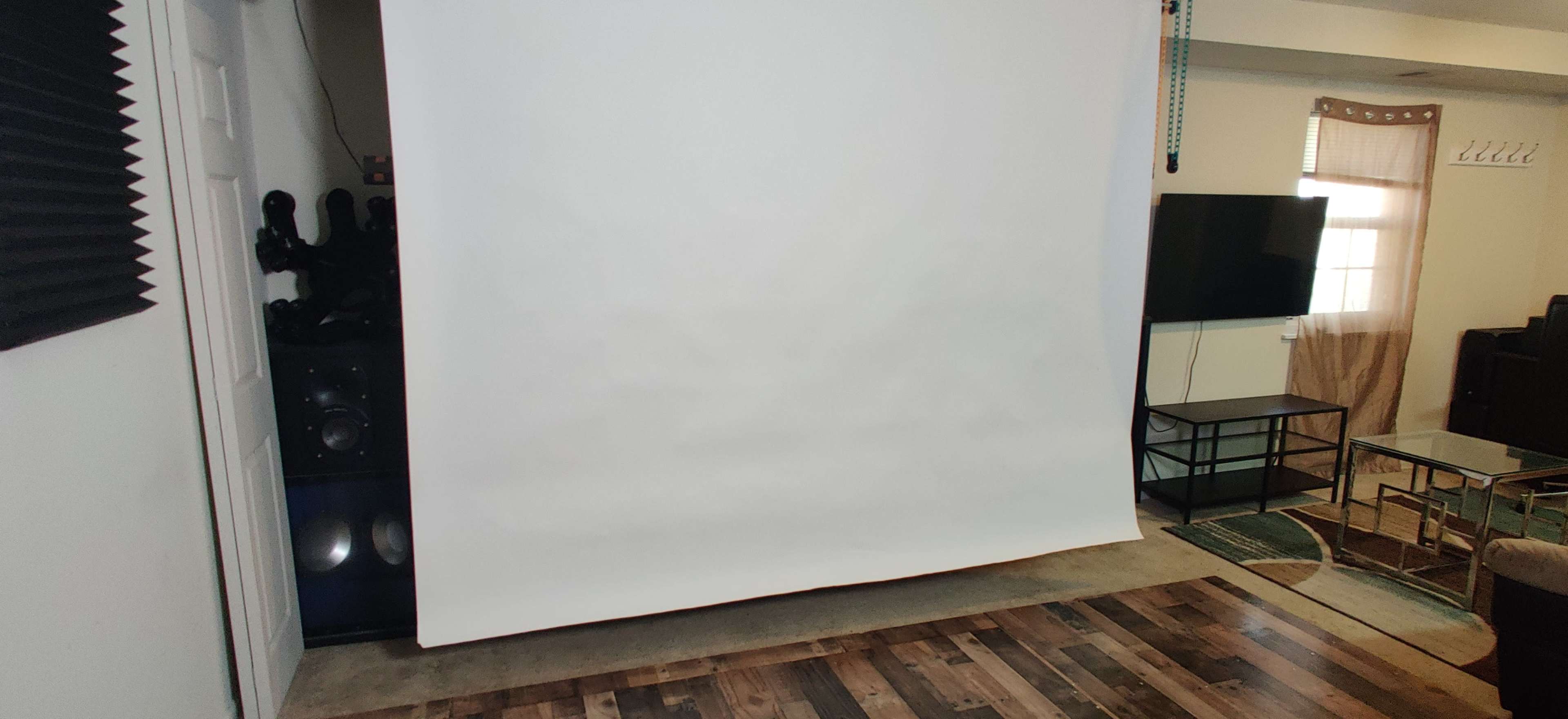 The image shows a room with a blank white projection screen mounted on the wall, a television on a stand, and a wooden floor.