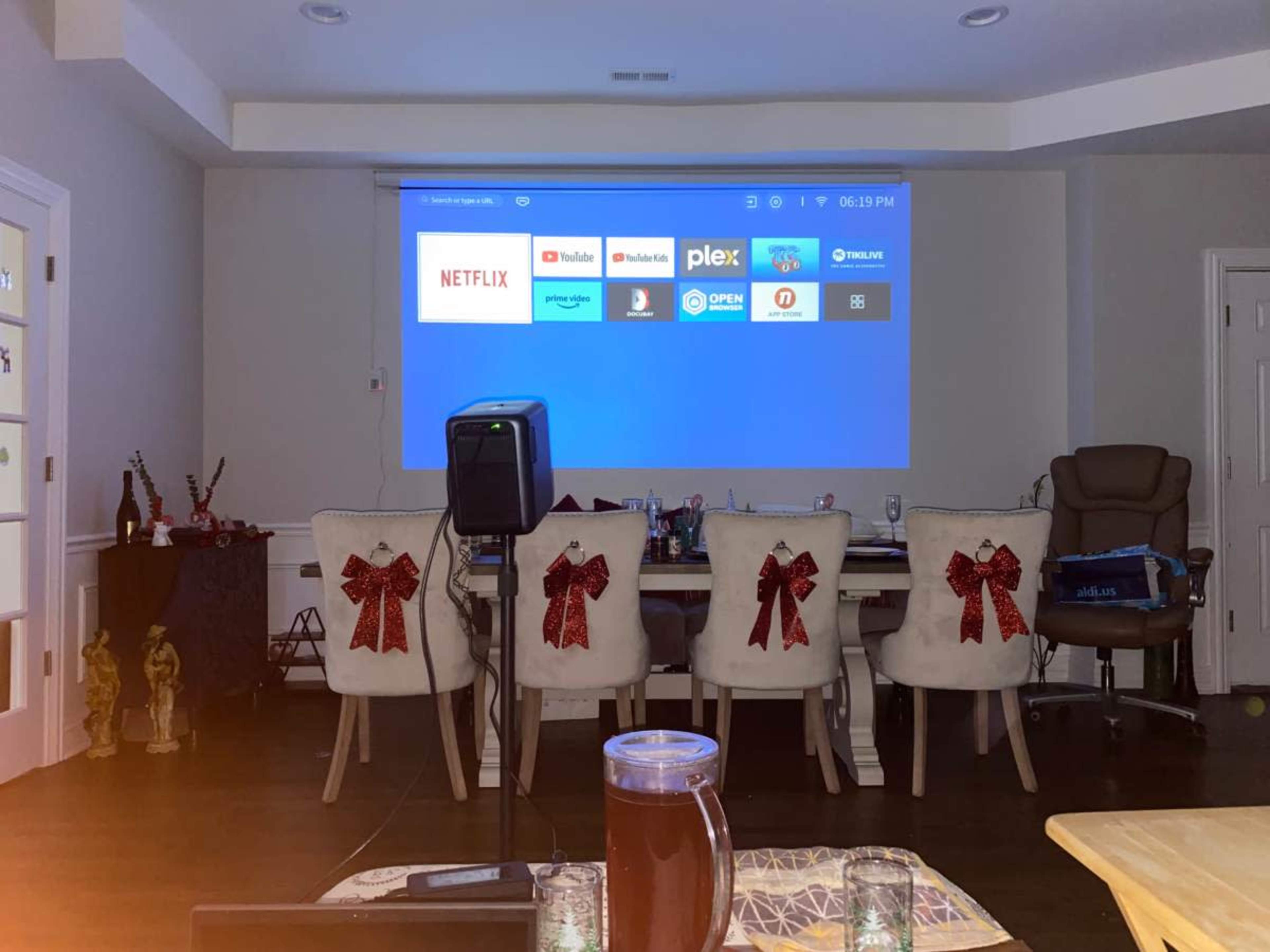 A large projector screen displays various app icons in a room with chairs adorned with red bows.