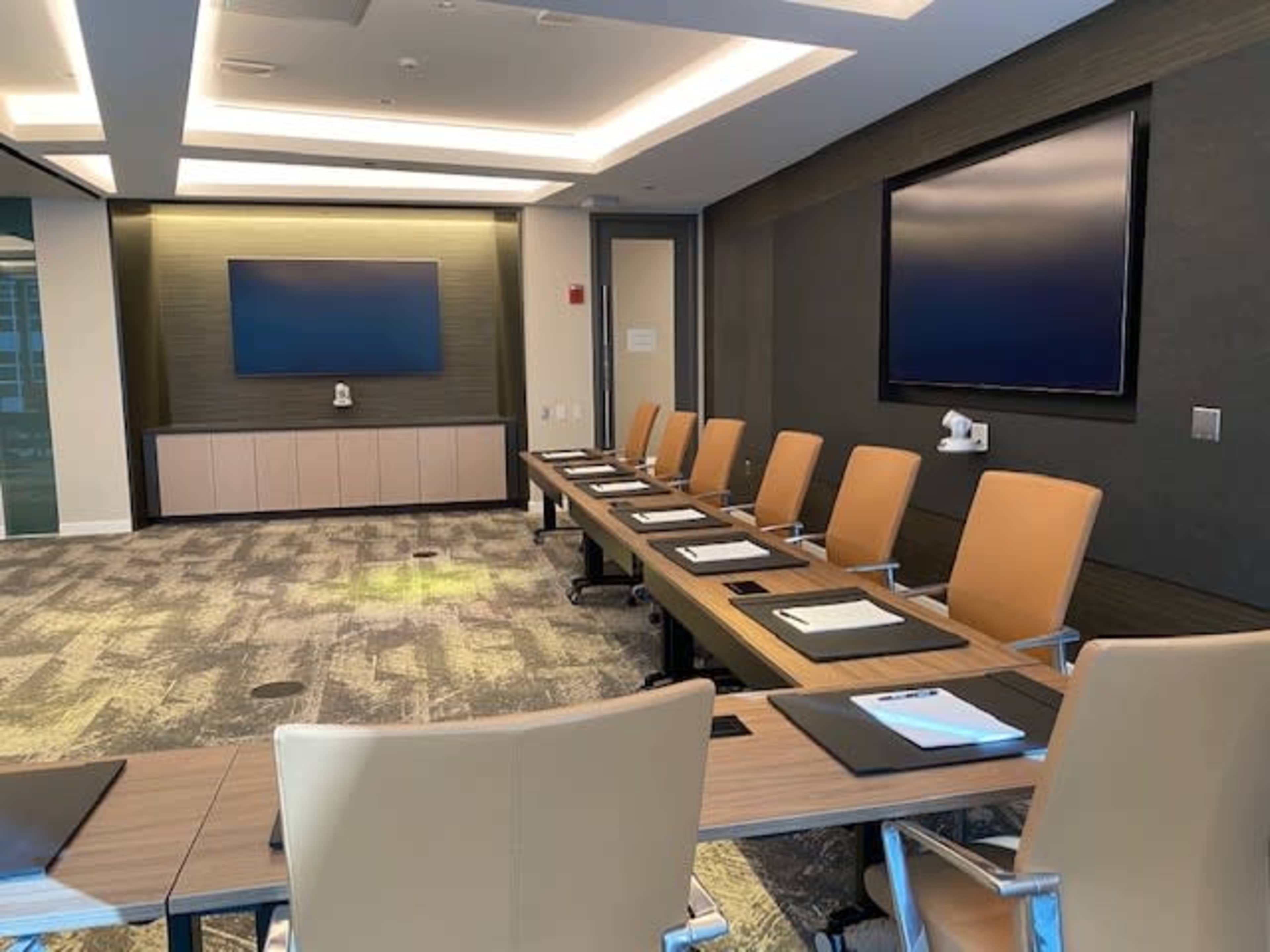 A modern conference room with a long table, several chairs, and two large screens mounted on the wall.