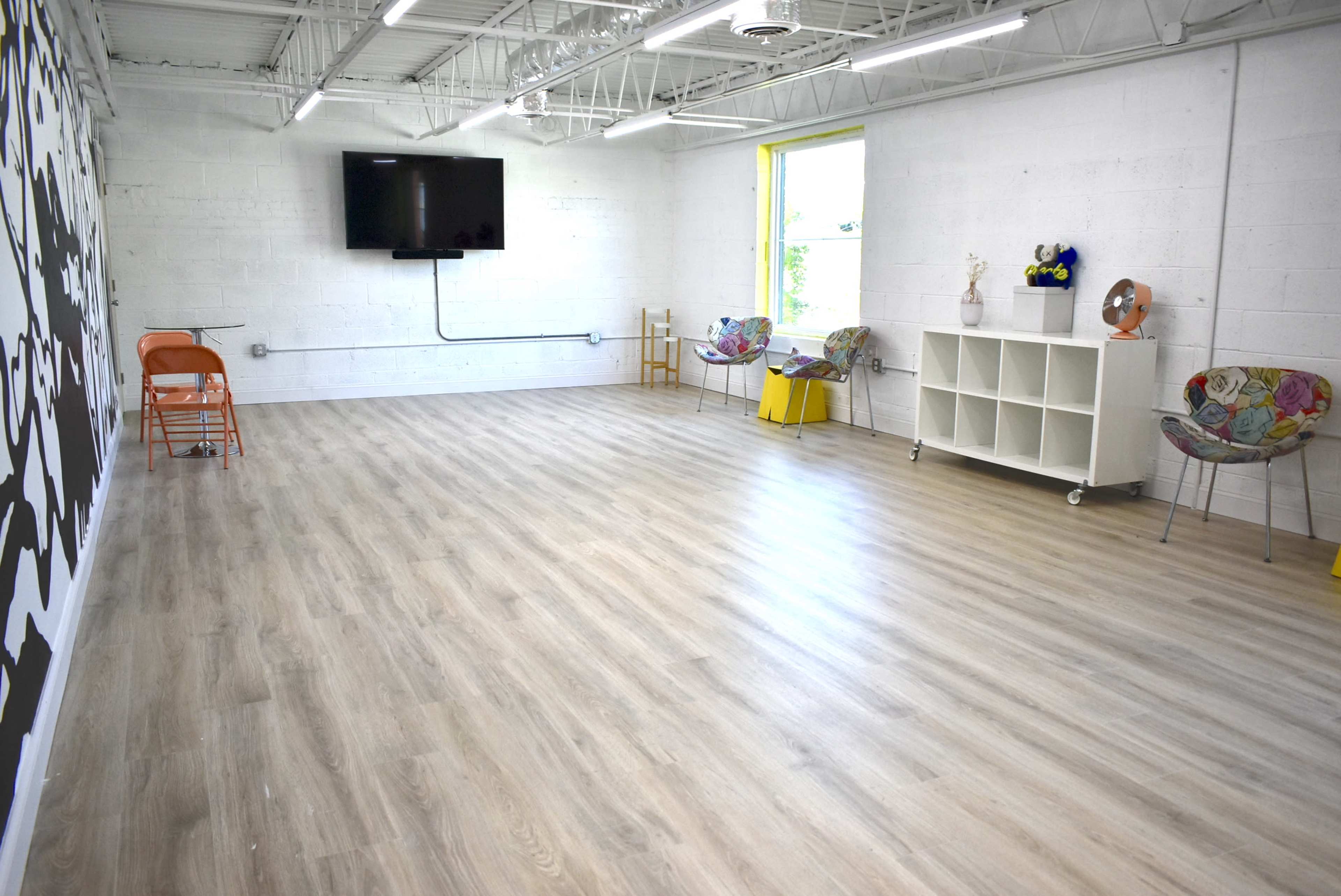 A spacious, well-lit room with light wooden flooring, a wall-mounted television, a window, and various furniture items including chairs and a shelving unit.