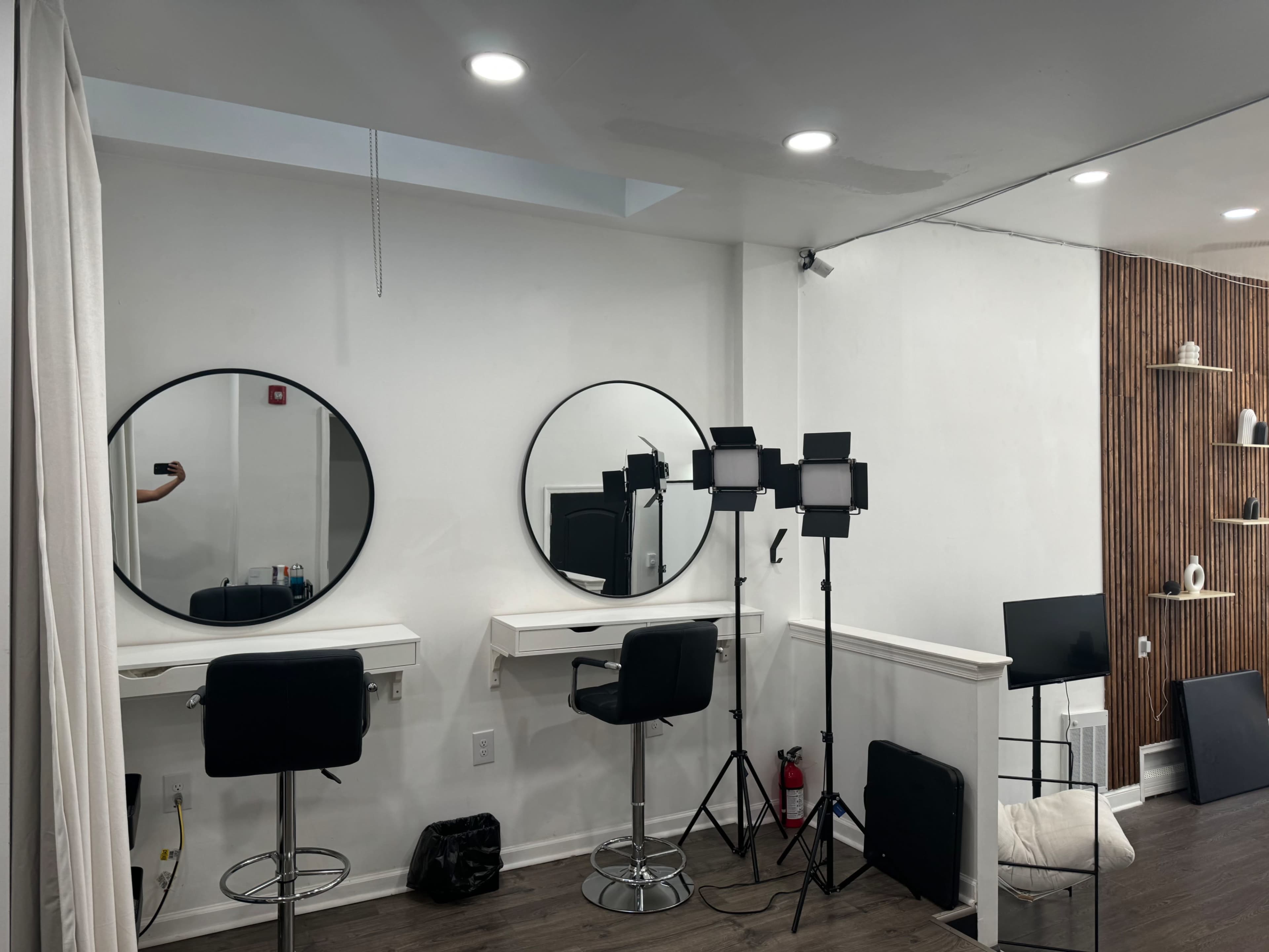 A modern makeup space with two round mirrors, two black bar stools, and studio lighting equipment set up against a white wall.