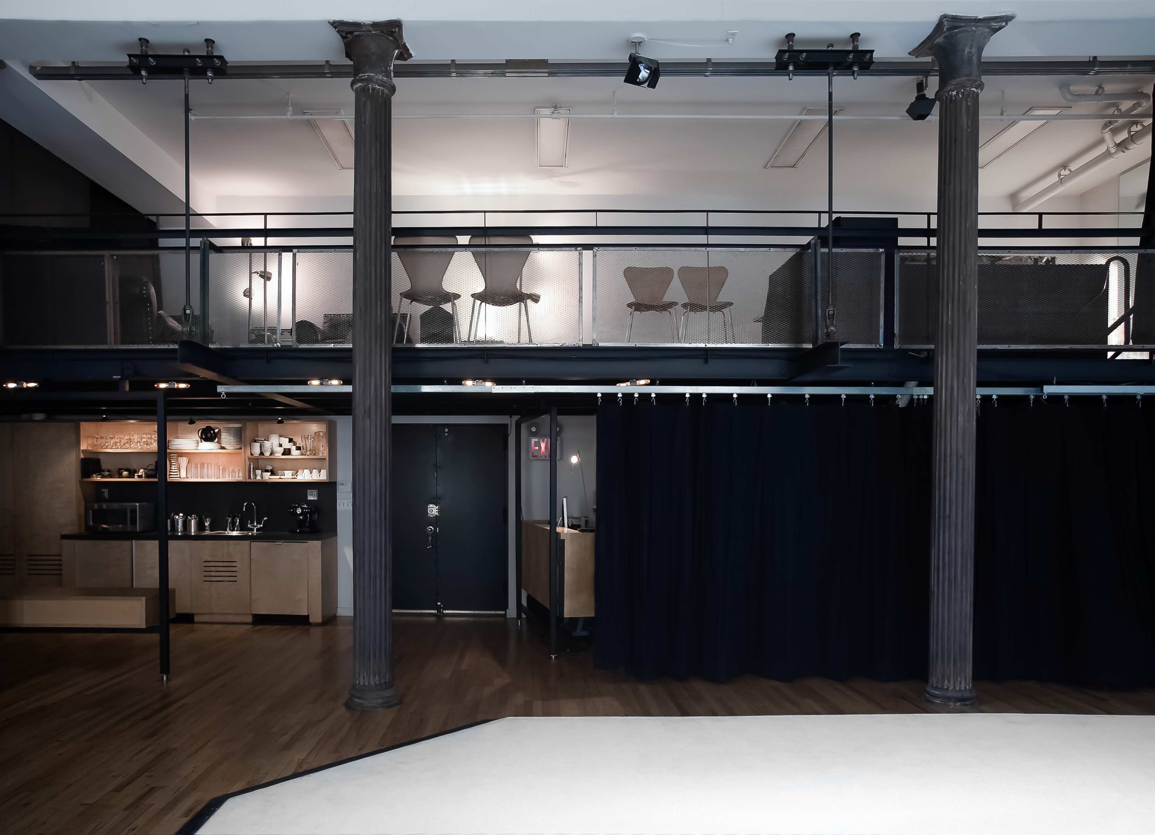 A modern interior space featuring a lower level with a bar and a black curtain, and an upper level with minimalist seating against glass railings.