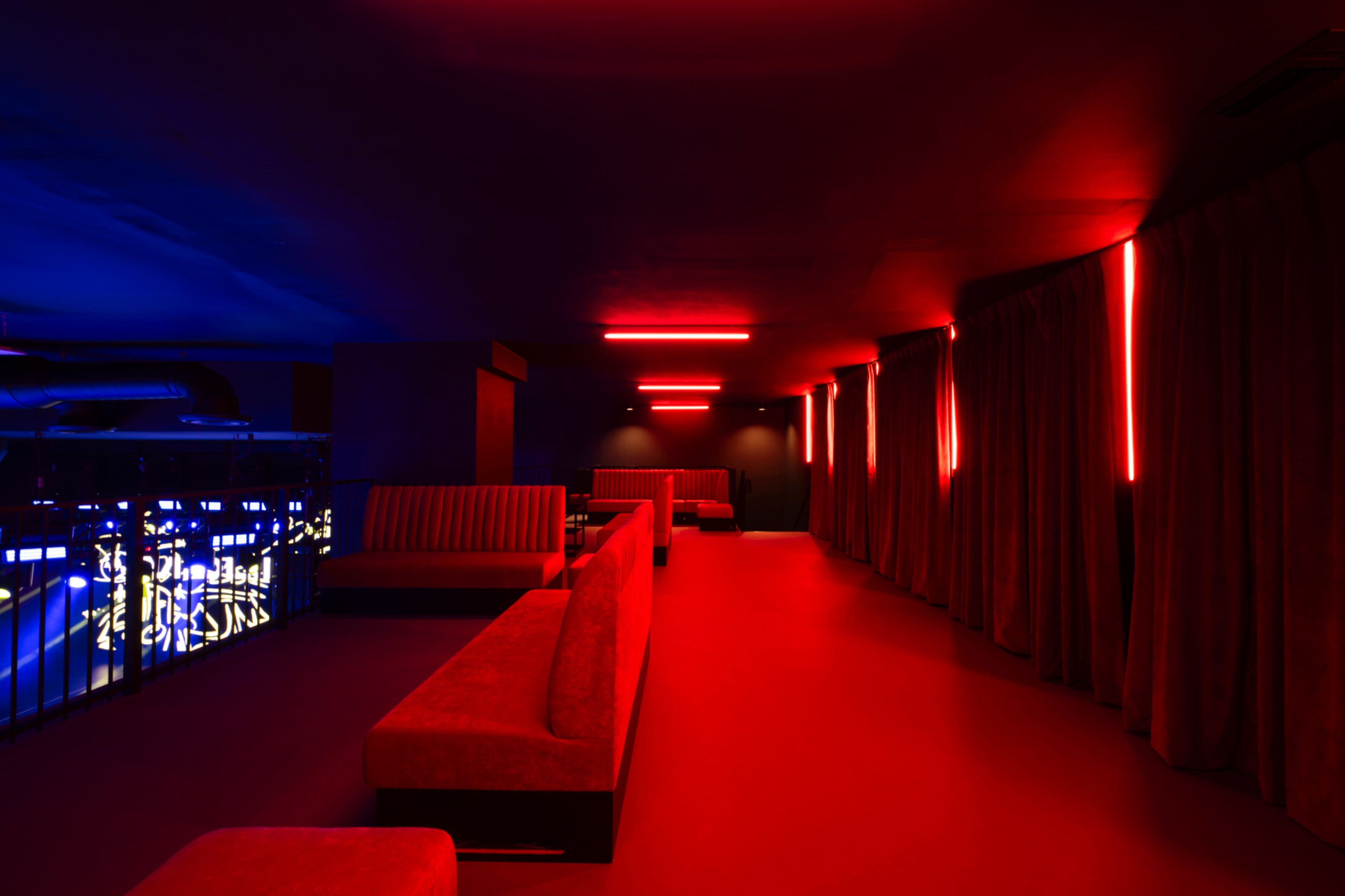 The image shows a dimly lit lounge featuring red neon lighting, dark walls, and plush red seating arranged along the walls.