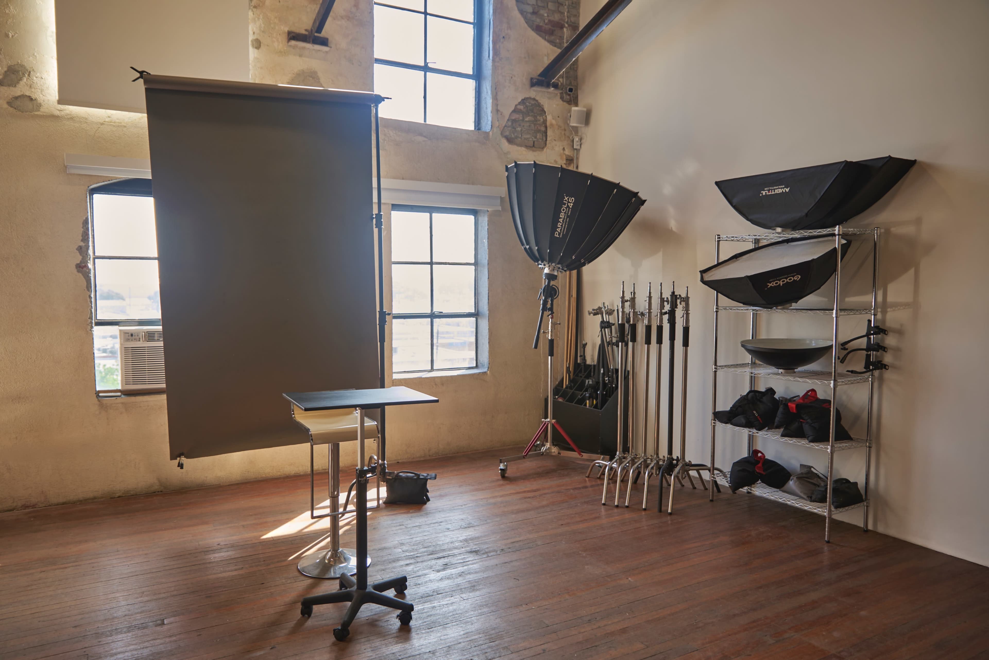 The image shows a photography studio with a gray backdrop, a table on wheels, various lighting equipment, and windows allowing natural light to enter.