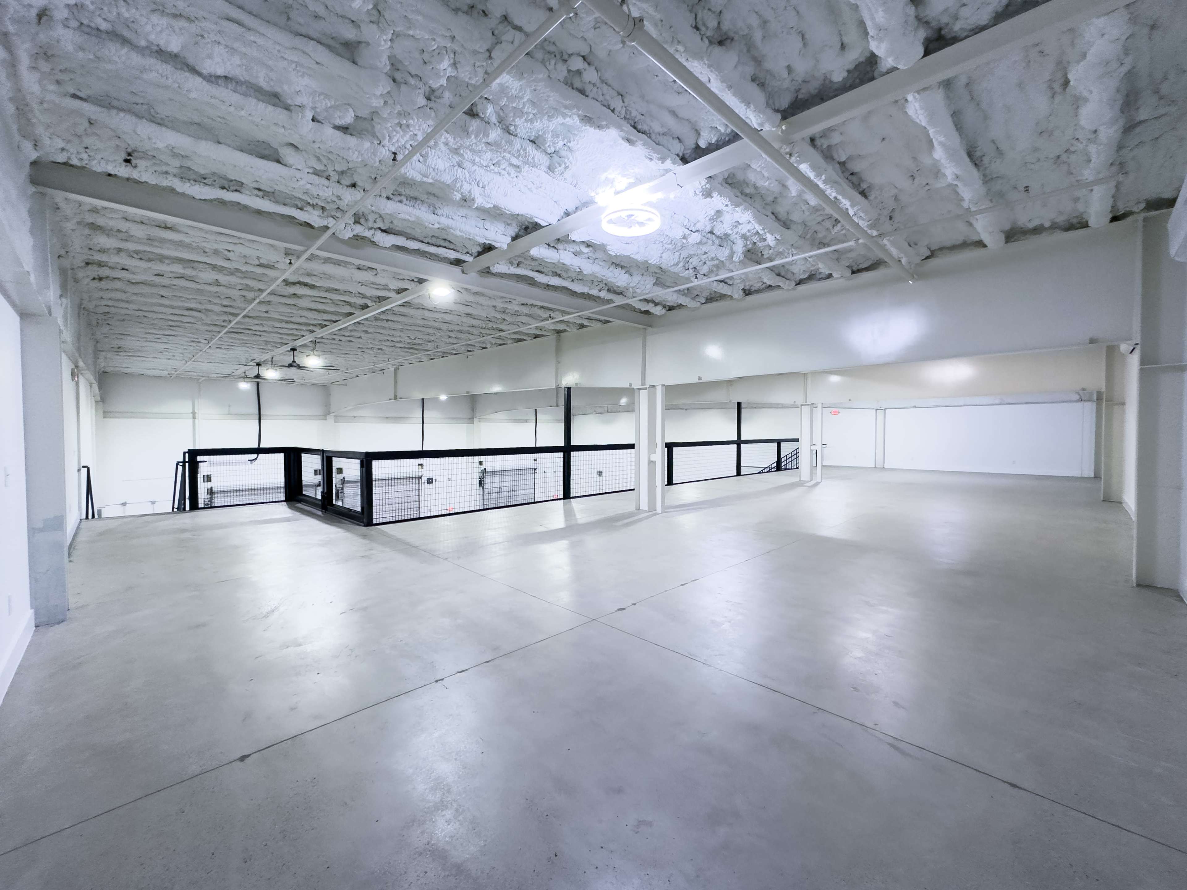 The image shows a spacious, empty interior of a building with exposed ceilings and concrete floors, featuring a balcony and metal railing.