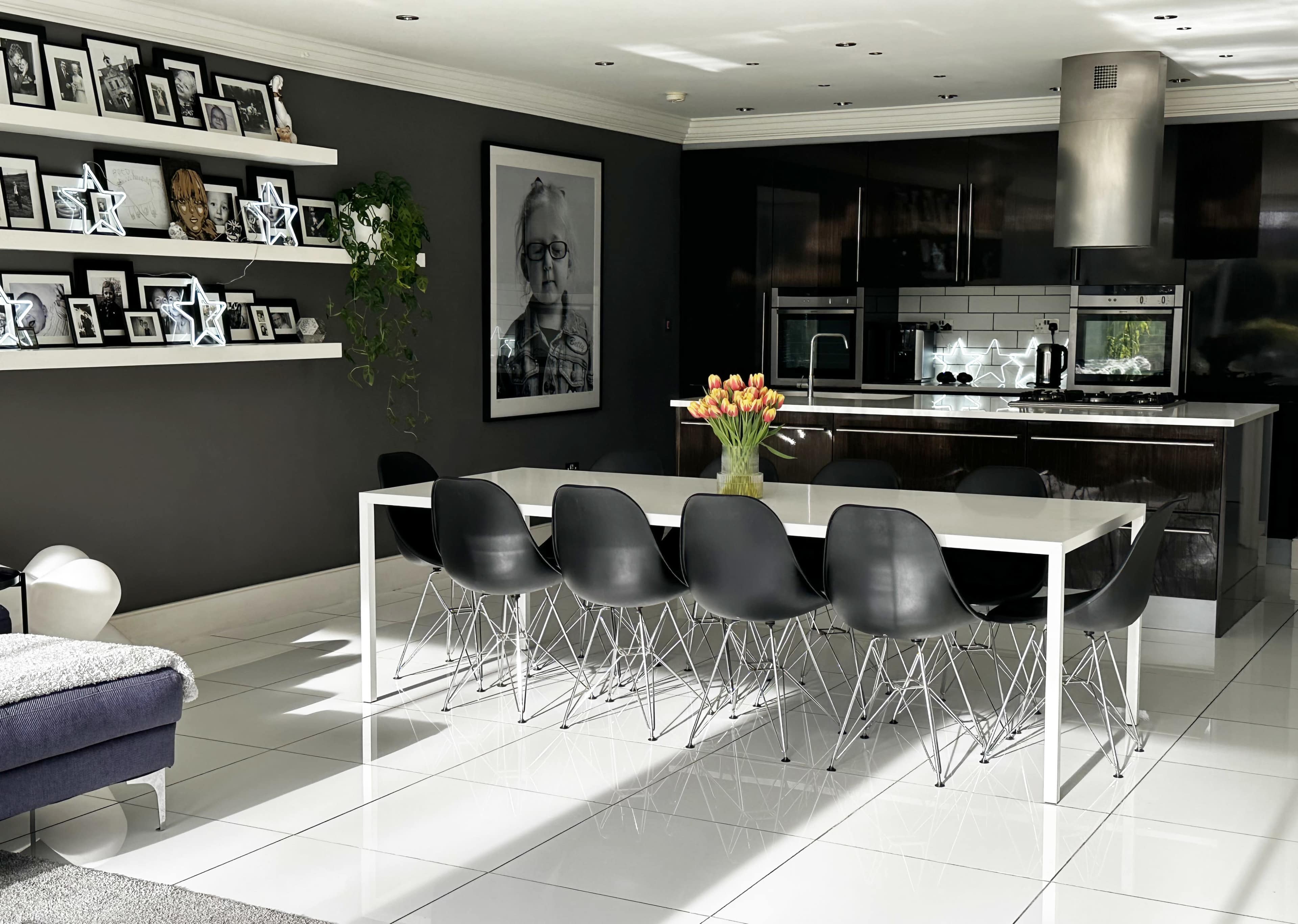 A modern dining area features a white table surrounded by black chairs, with a wall of framed photos and a kitchen in the background.