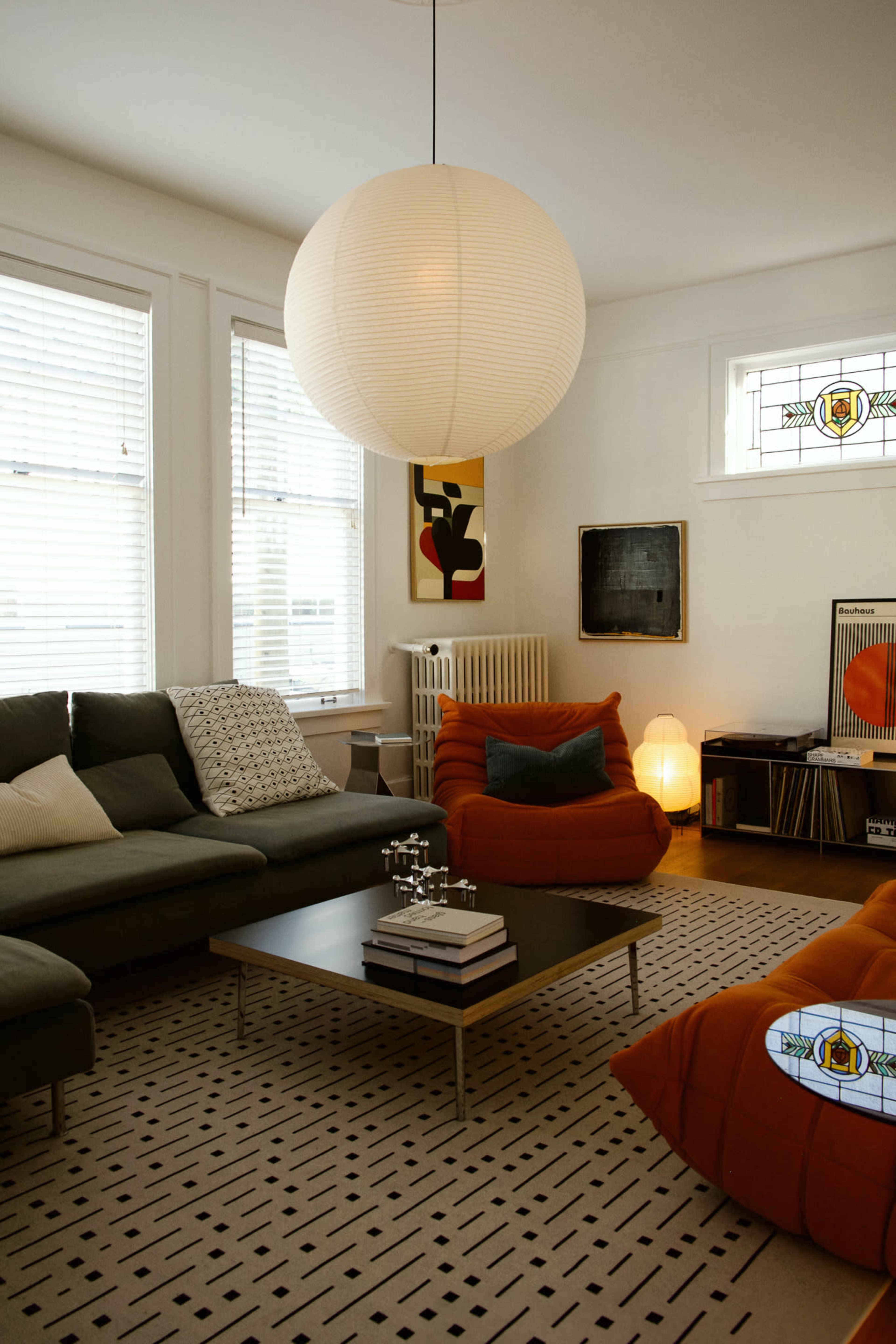 The image shows a modern living room featuring a gray sectional sofa, orange armchair, a coffee table with books, and large windows adorned with blinds.