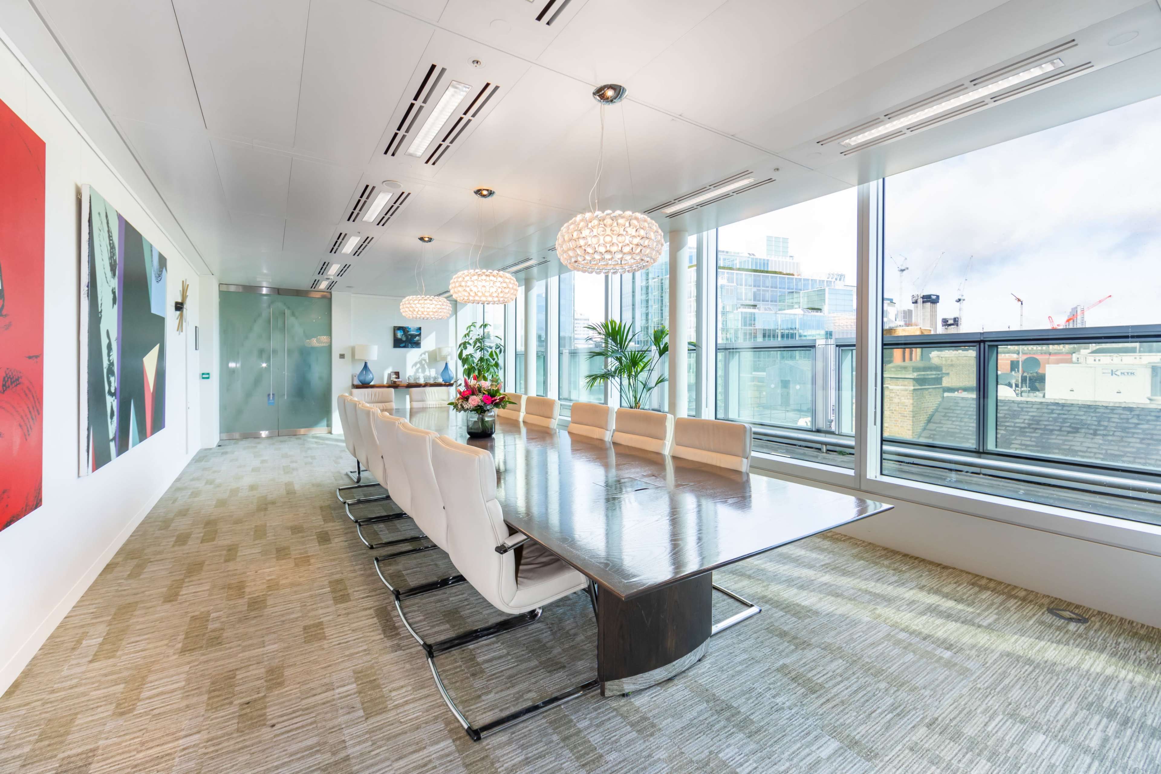 A modern conference room features a large wooden table surrounded by white leather chairs, with floor-to-ceiling windows and contemporary lighting.