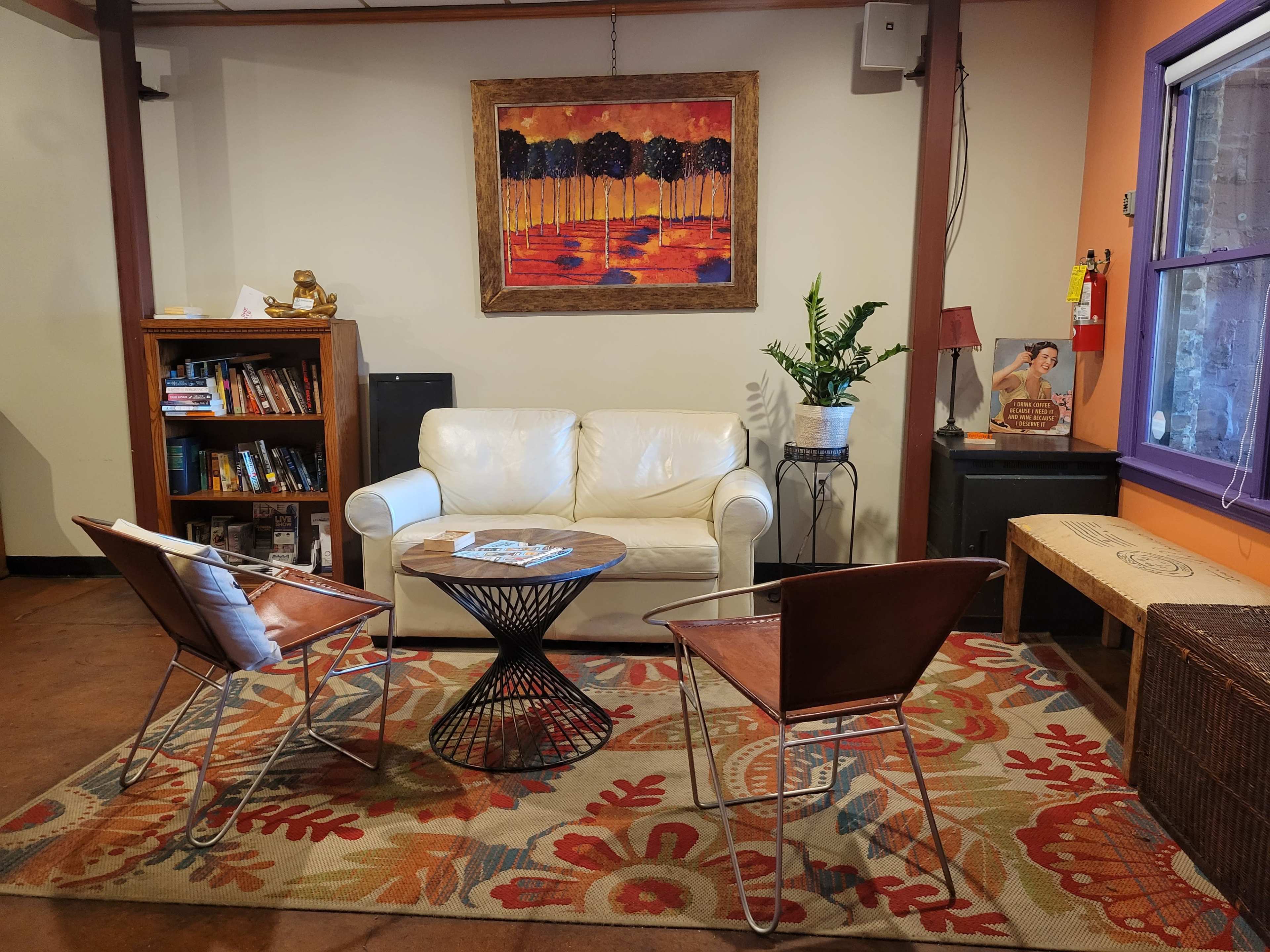 A cozy lounge area with a white sofa, two brown chairs, a round black coffee table, a colorful rug, and a bookshelf against the wall.