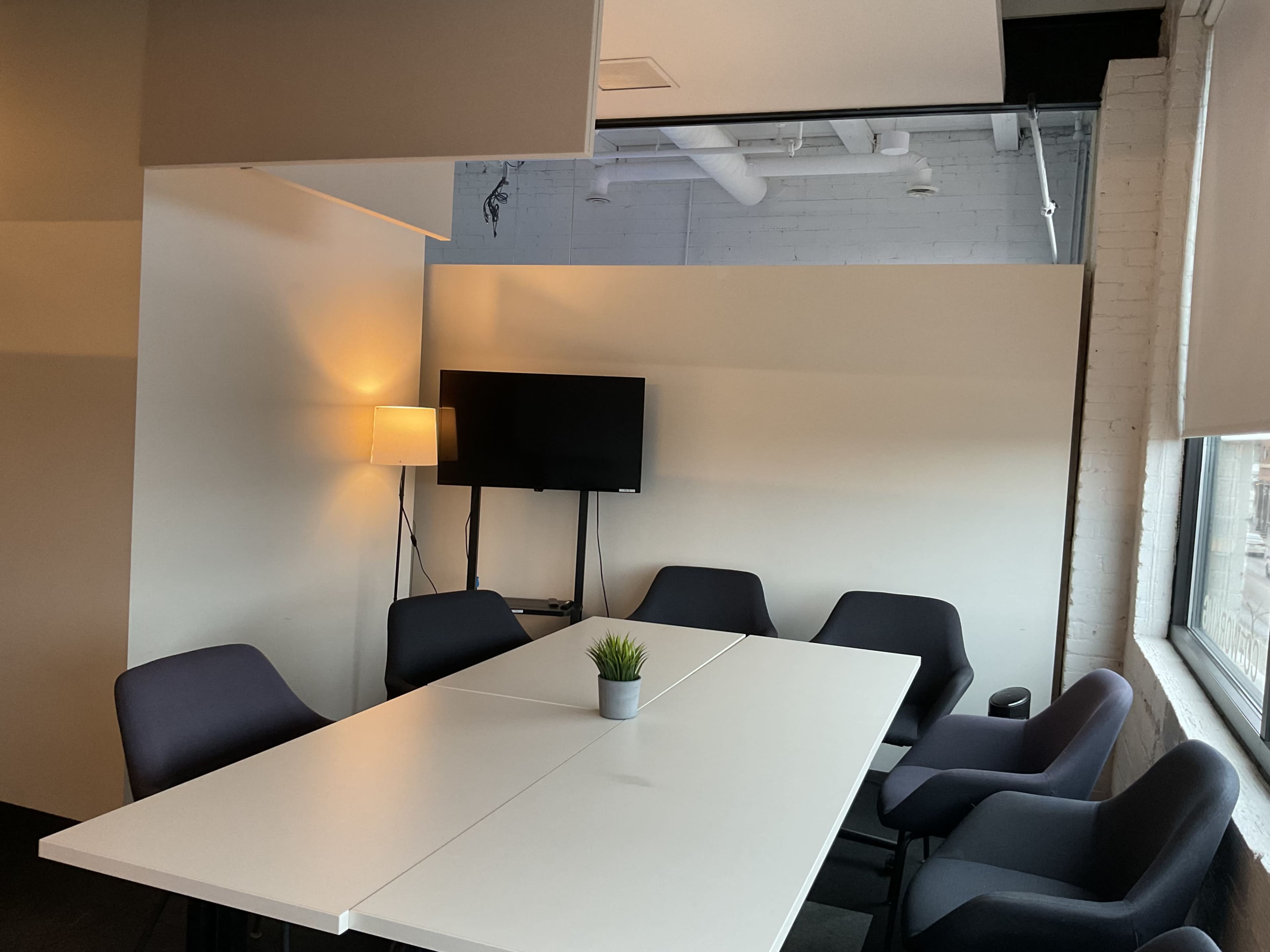 The image shows a conference room with a large white table, several chairs, a lamp, and a television mounted on the wall.