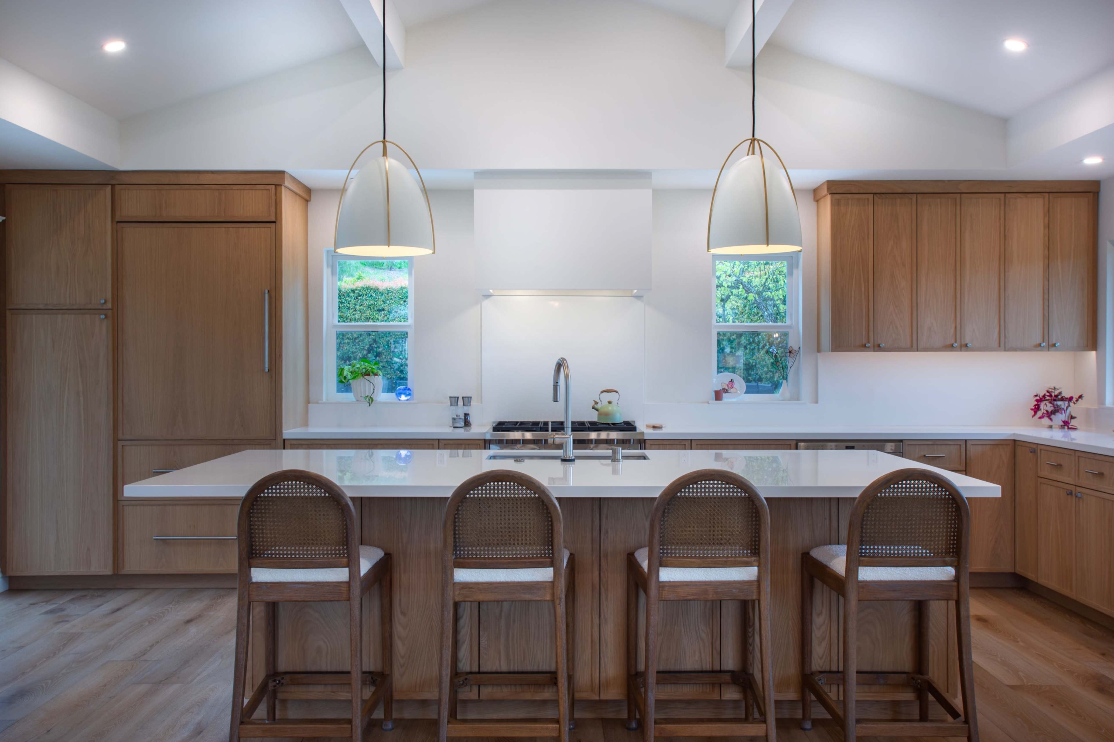 A modern kitchen features a large island with four bar stools, wooden cabinetry, and large windows letting in natural light.