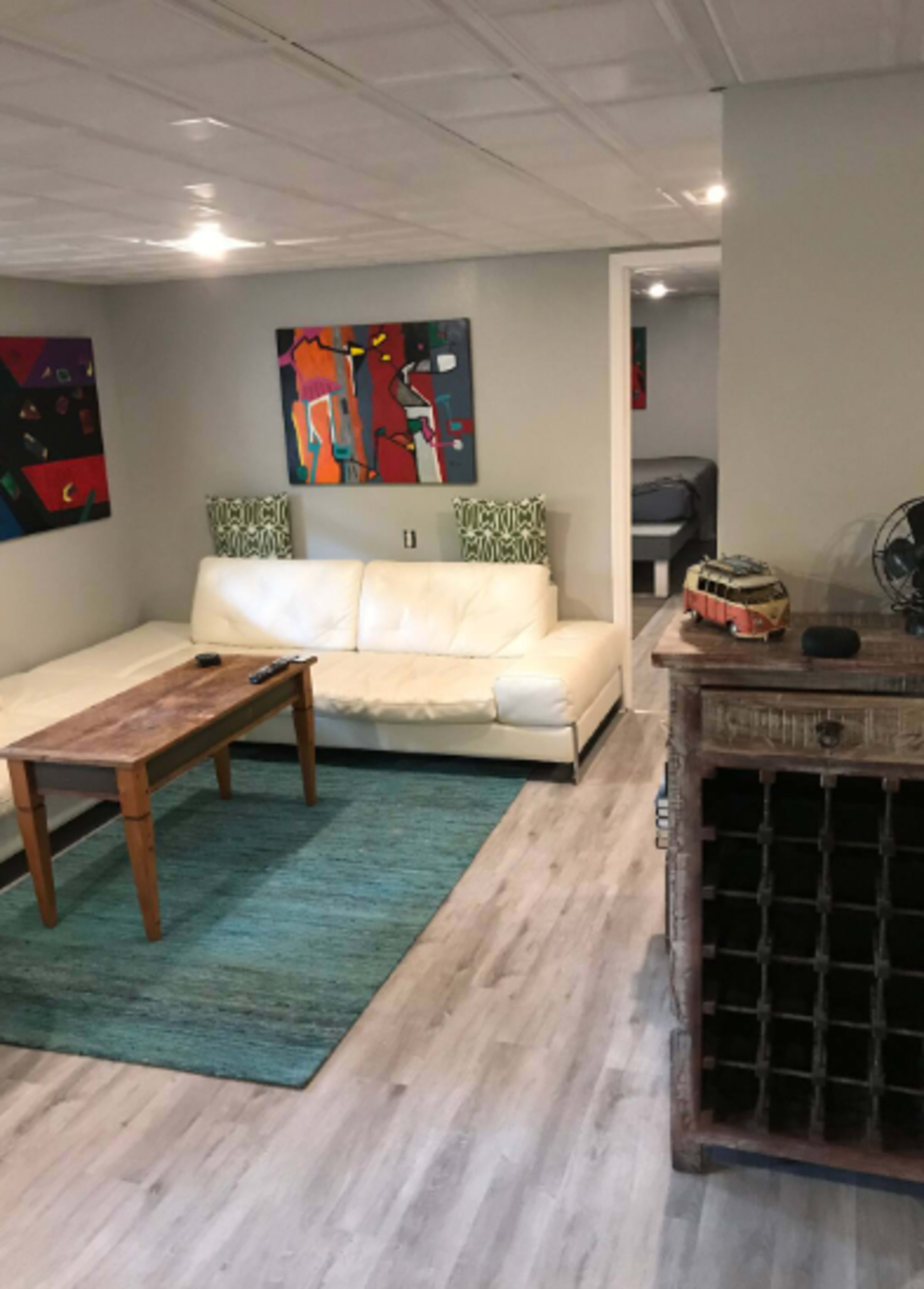 The image shows a cozy living room with a white sofa, a wooden coffee table, and a textured rug in front, complemented by colorful artworks on the walls.