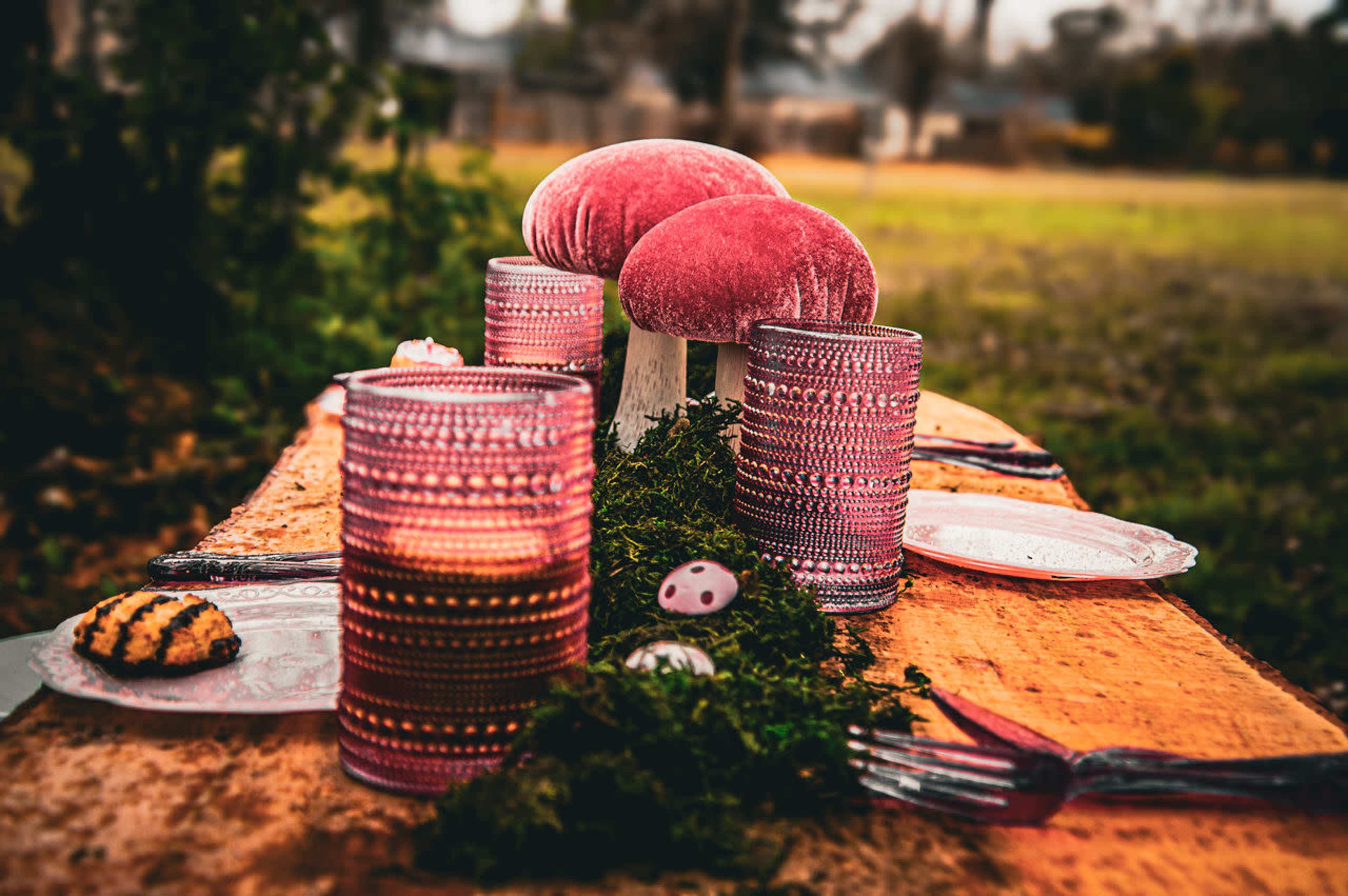 A rustic wooden table is set outdoors with pink patterned glassware, decorative mushrooms, and plates, surrounded by greenery.