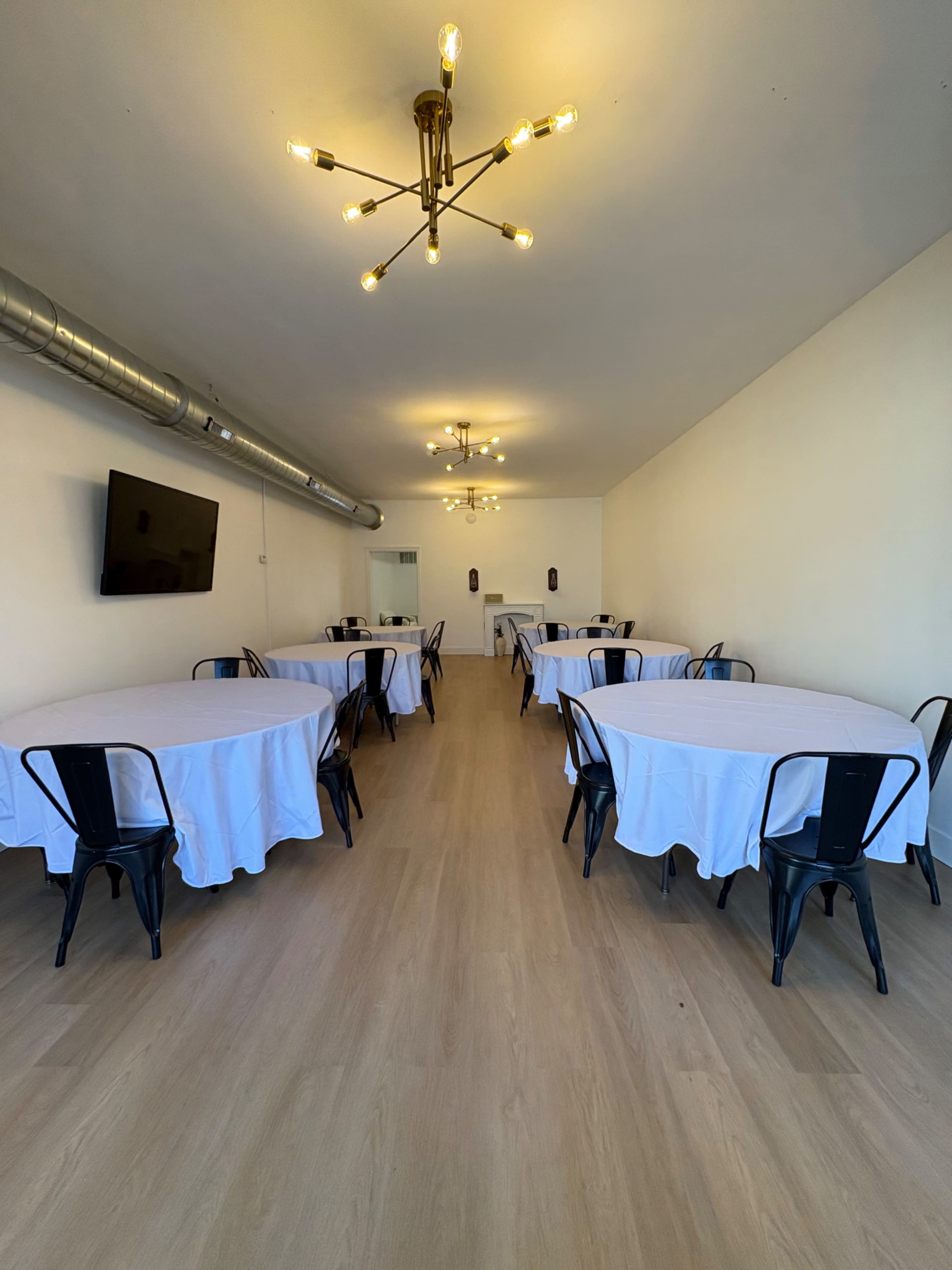 The image shows a spacious room with several round tables covered in white tablecloths and black chairs, illuminated by modern light fixtures.