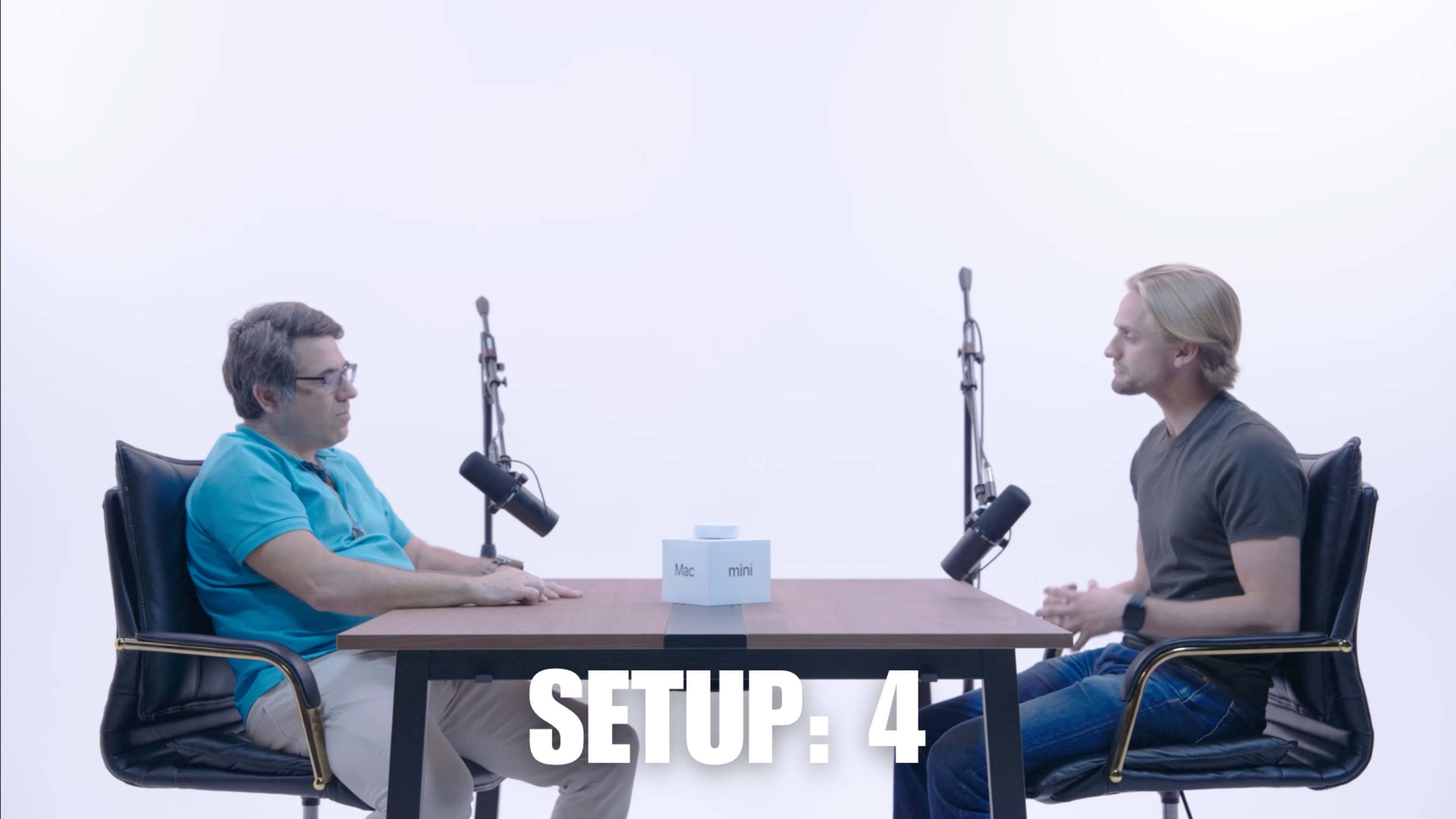 Two men are seated at a table with microphones in a bright, minimalistic setting.
