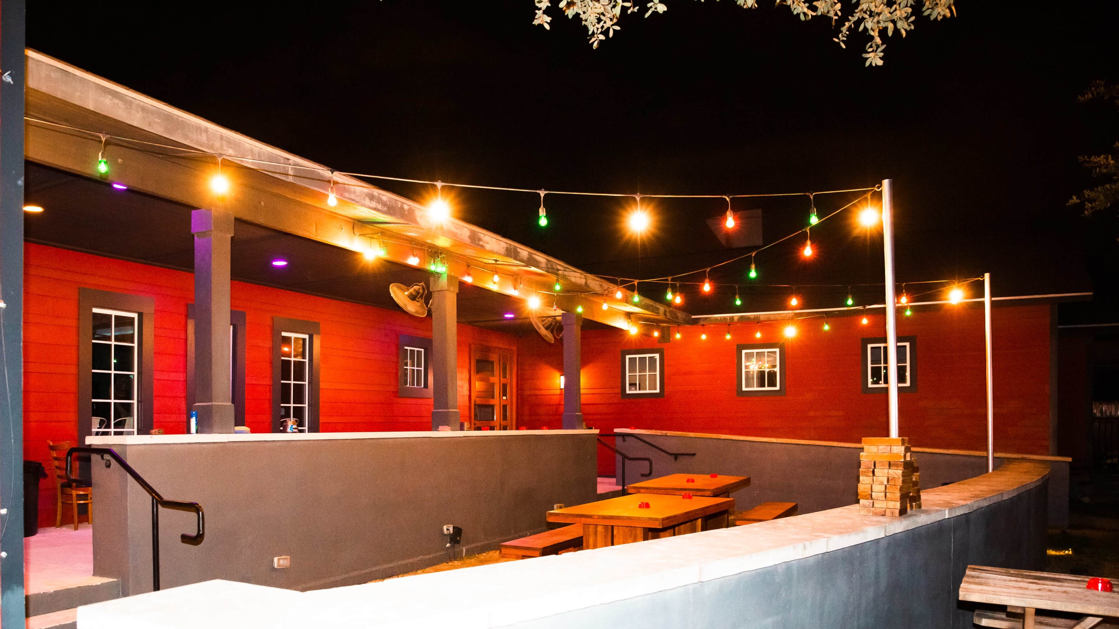 The image shows a brightly lit outdoor patio area with string lights illuminating wooden tables and a red-paneled building in the background.