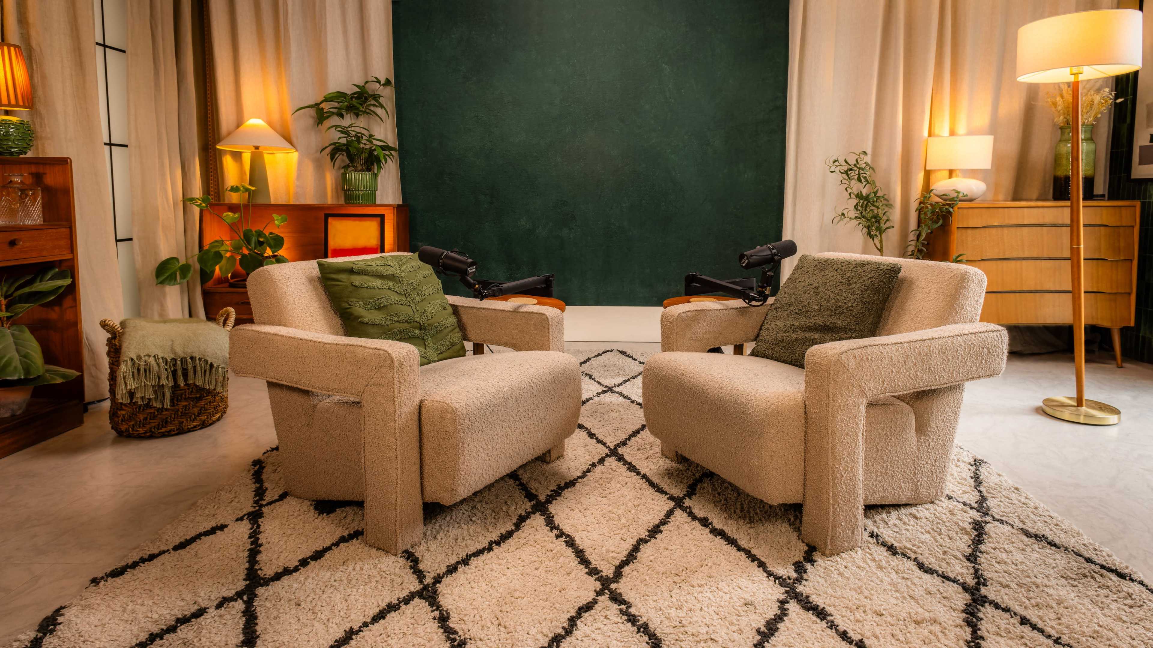 The image shows two beige armchairs facing each other on a patterned rug in a warmly lit room with plants and lamps.