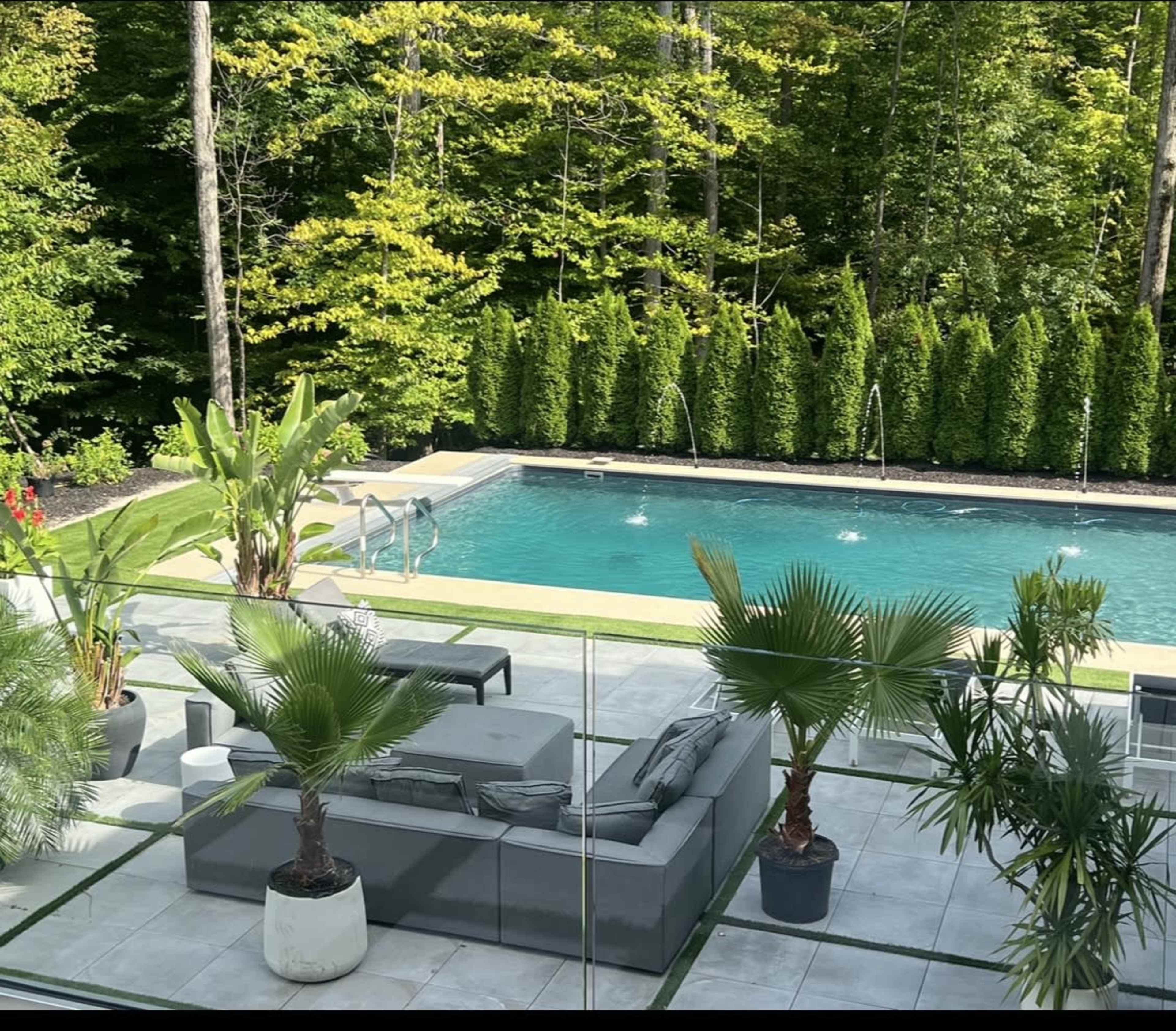 A modern outdoor patio features a gray sectional sofa and potted palm plants, overlooking a swimming pool surrounded by lush greenery.