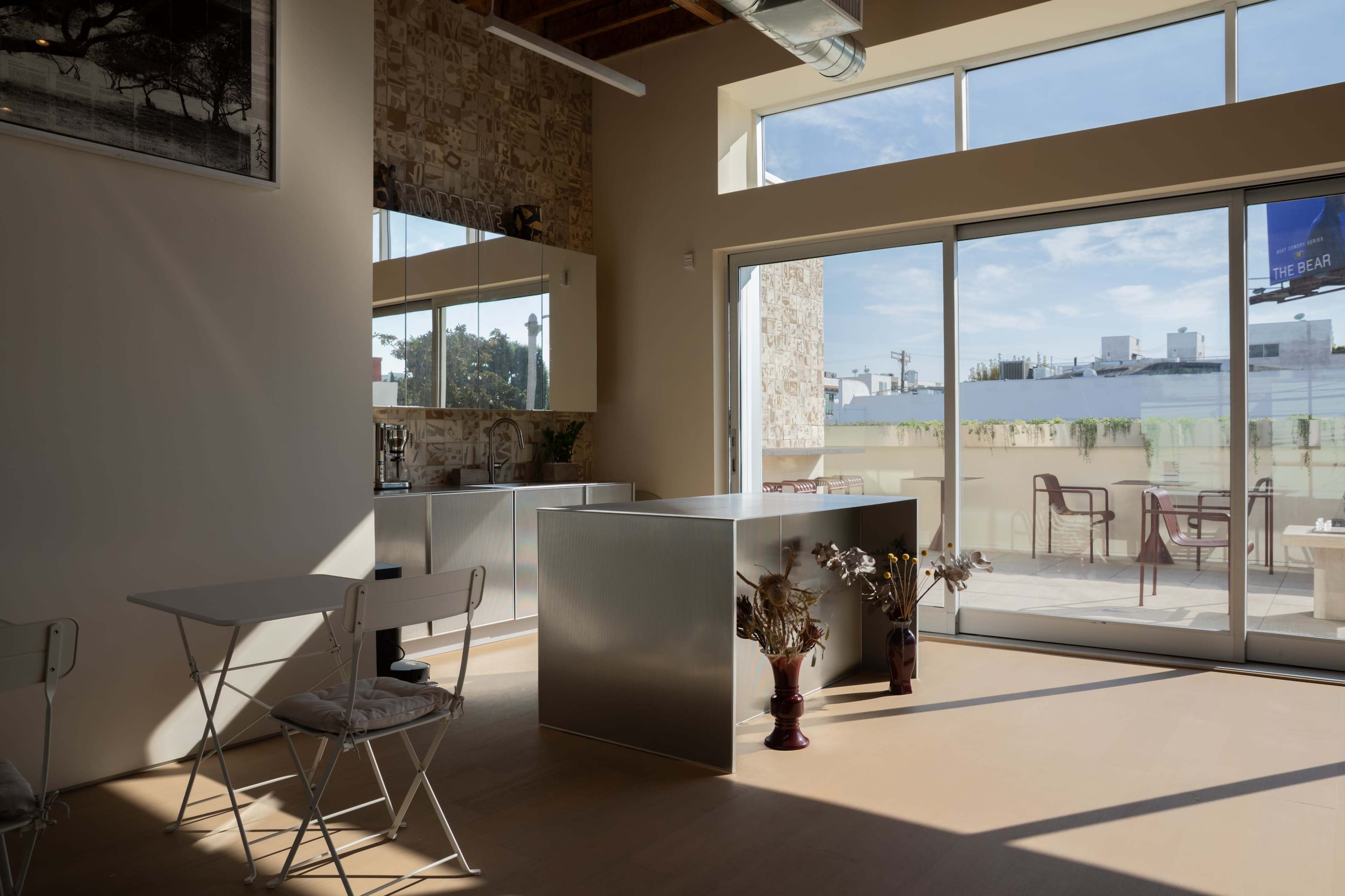 The image shows a modern, minimalist interior featuring a silver kitchen island, a small white folding table with chairs, and large windows that allow natural light to illuminate the space.