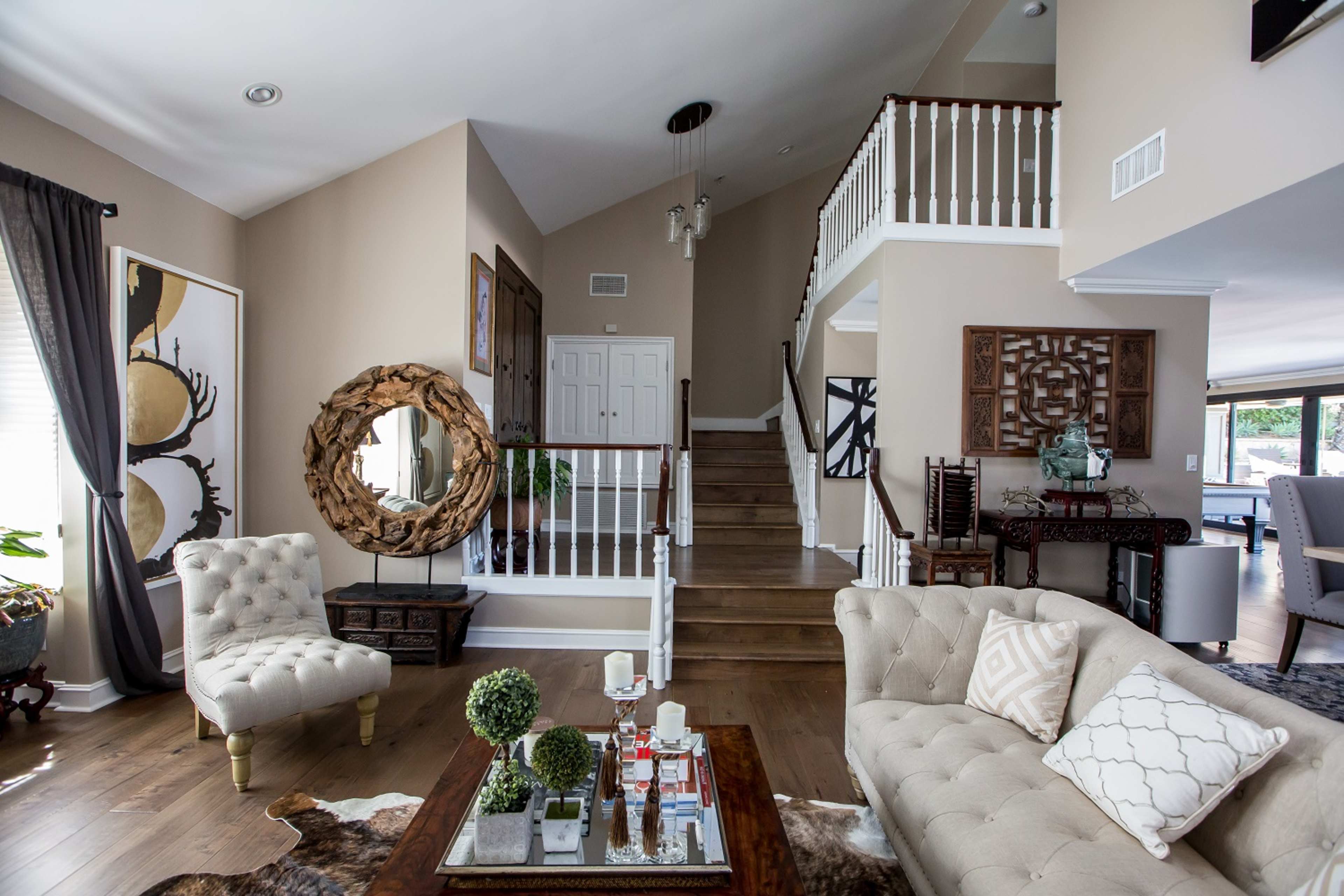 The image shows a spacious living room with light-colored walls, a staircase leading to an upper level, and a circular mirror on the wall beside comfortable seating and a coffee table.