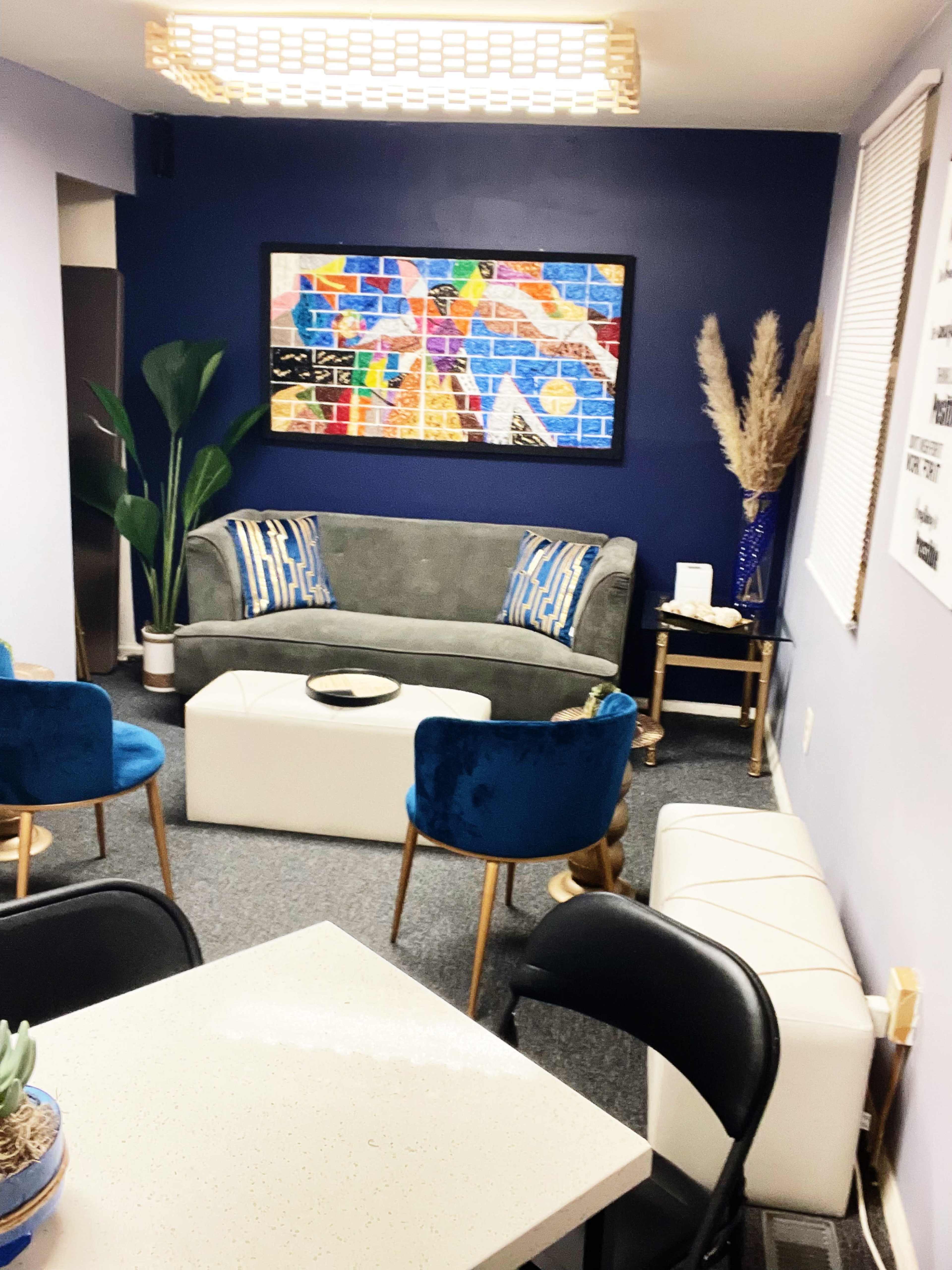 A cozy sitting area with a gray sofa, blue accent chairs, and a decorative wall art piece, all set in a room with blue walls and a light-colored table.