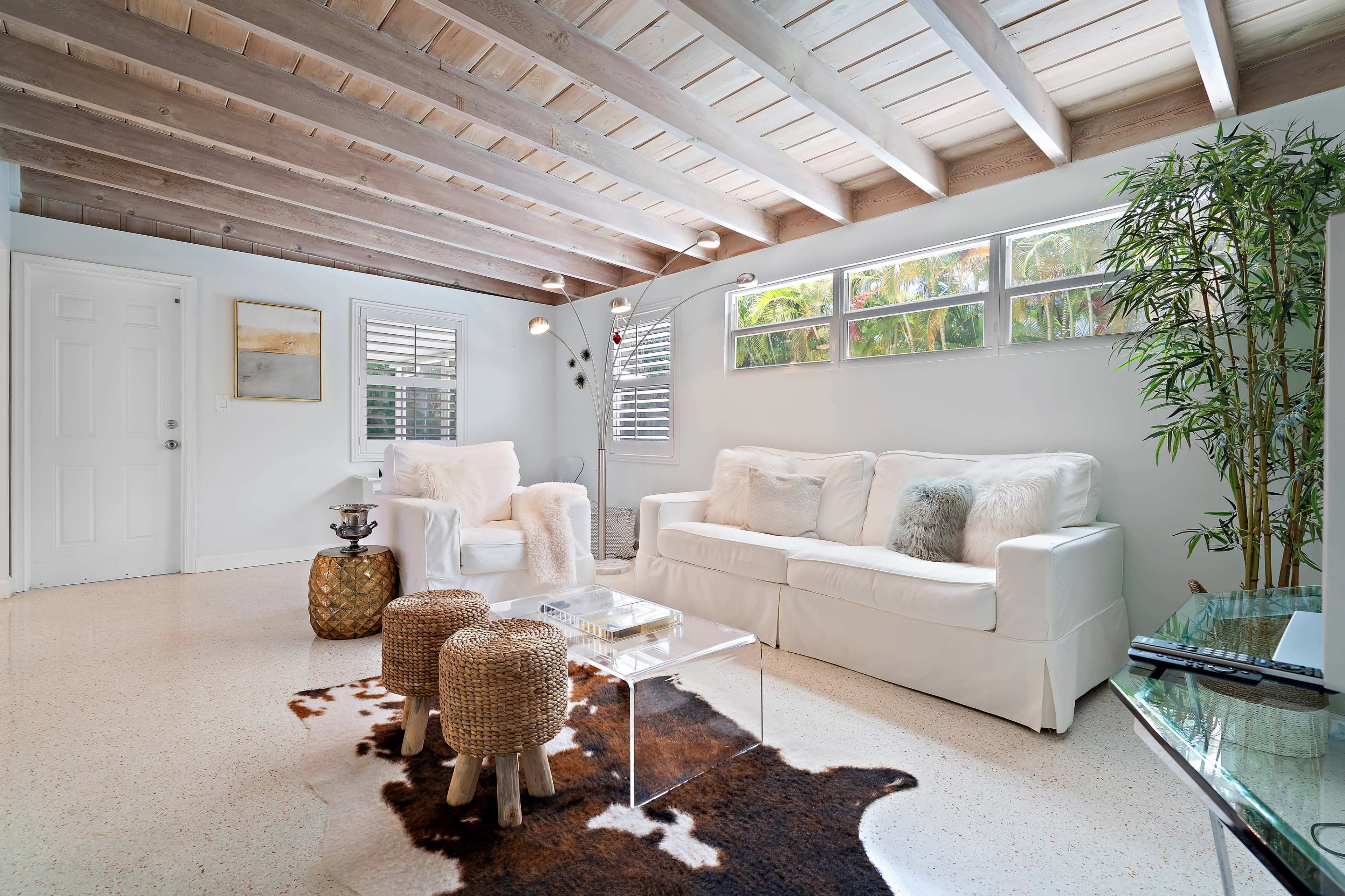 A bright living room with a white sofa and chairs, a glass coffee table, and wooden ceiling beams, complemented by a small plant.