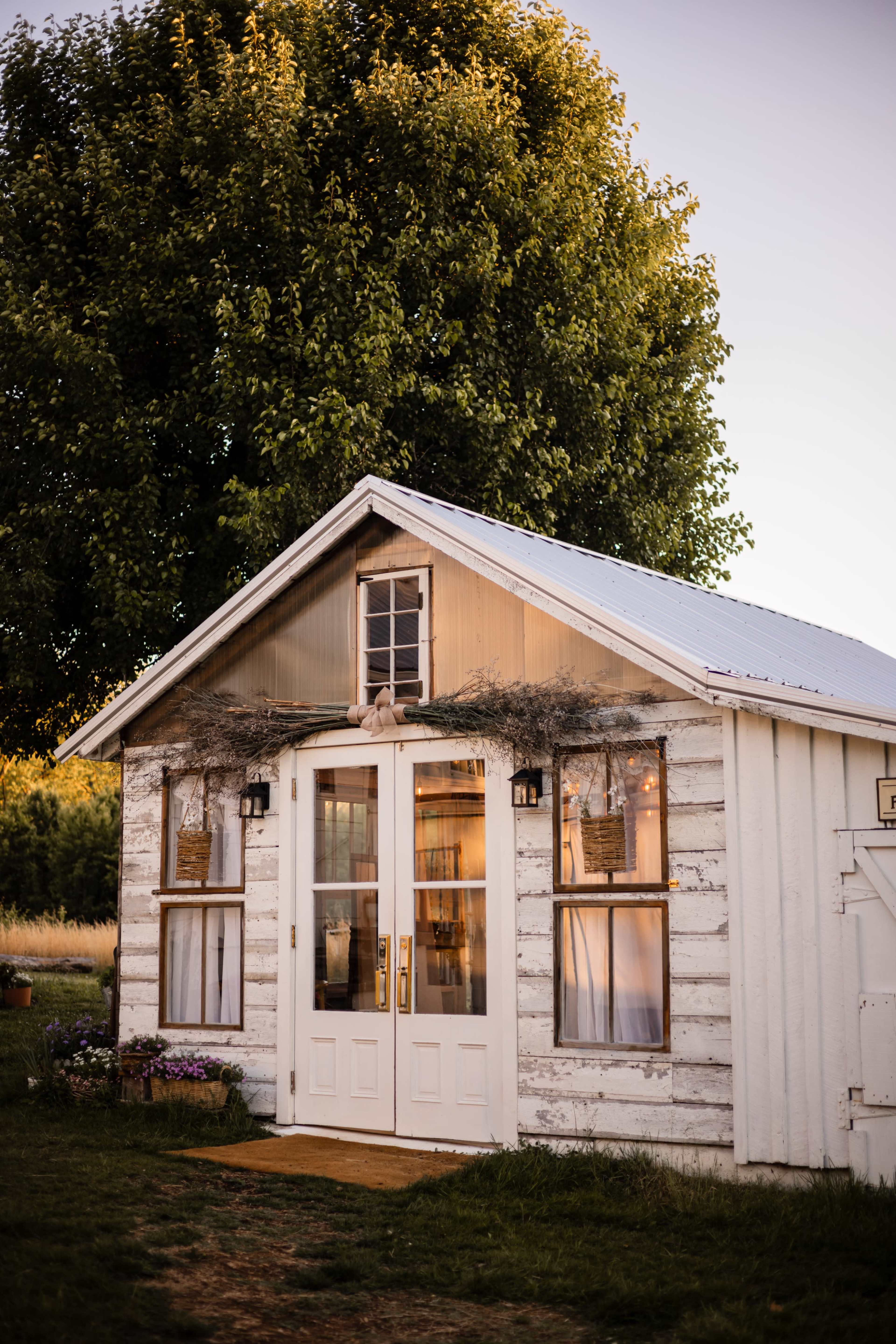The image shows a small, rustic wooden cabin with a porch and large tree nearby, set in a grassy area during sunset.