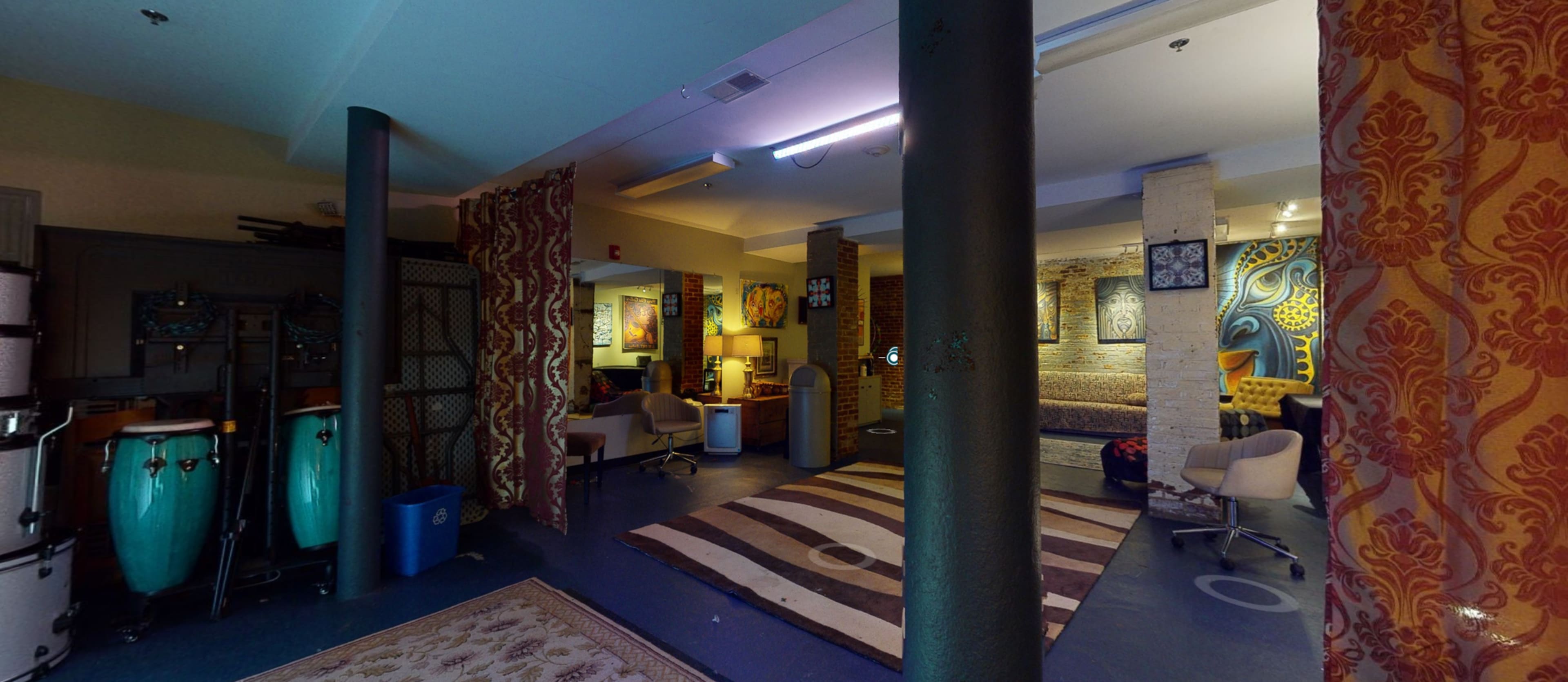 The image shows a spacious room with musical instruments, a decorative rug, and various artworks on the walls, separated by columns and curtains.