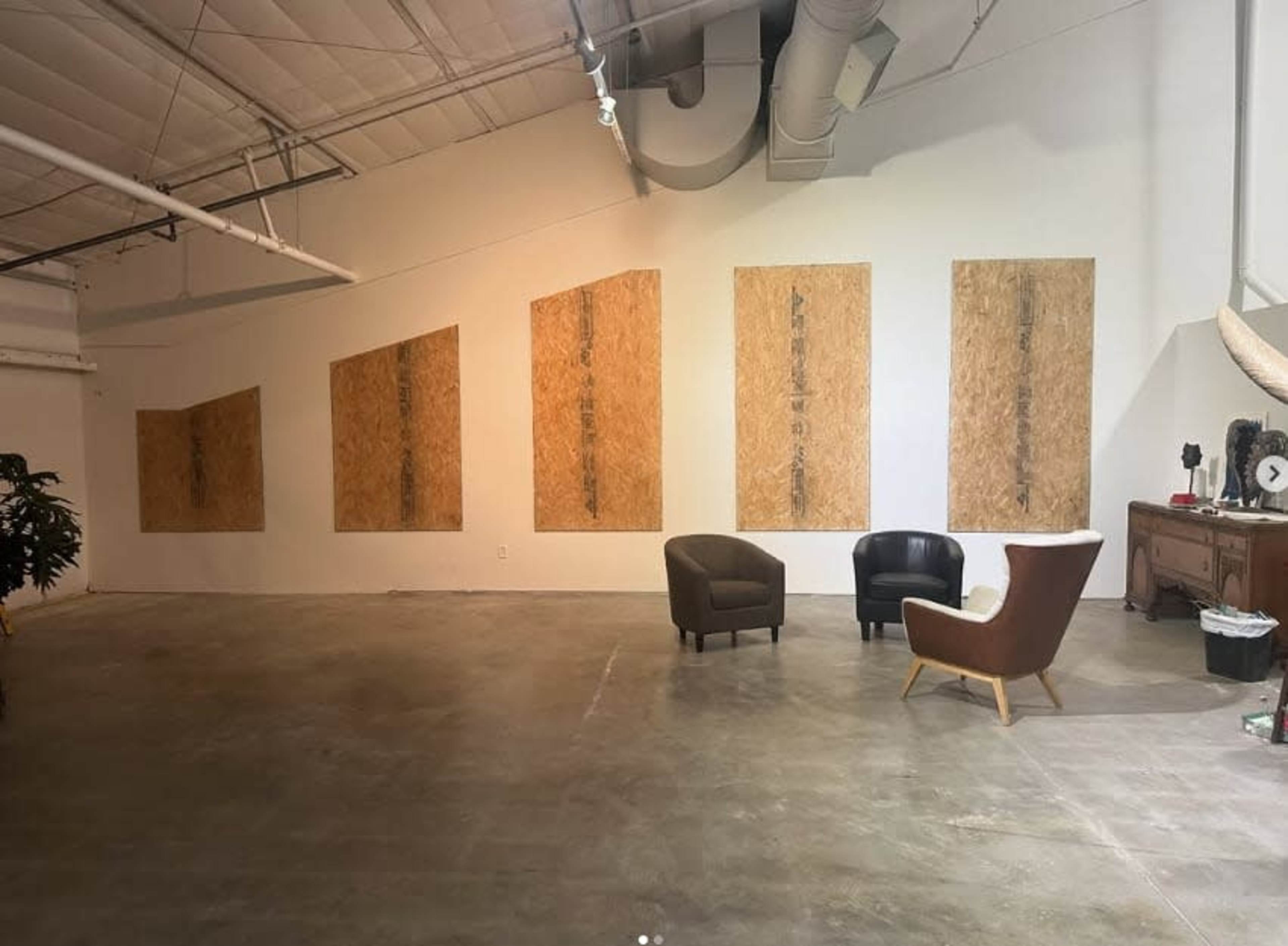The image shows an industrial-style room with concrete floors, featuring six large, boarded-up openings on one wall and two armchairs positioned nearby.
