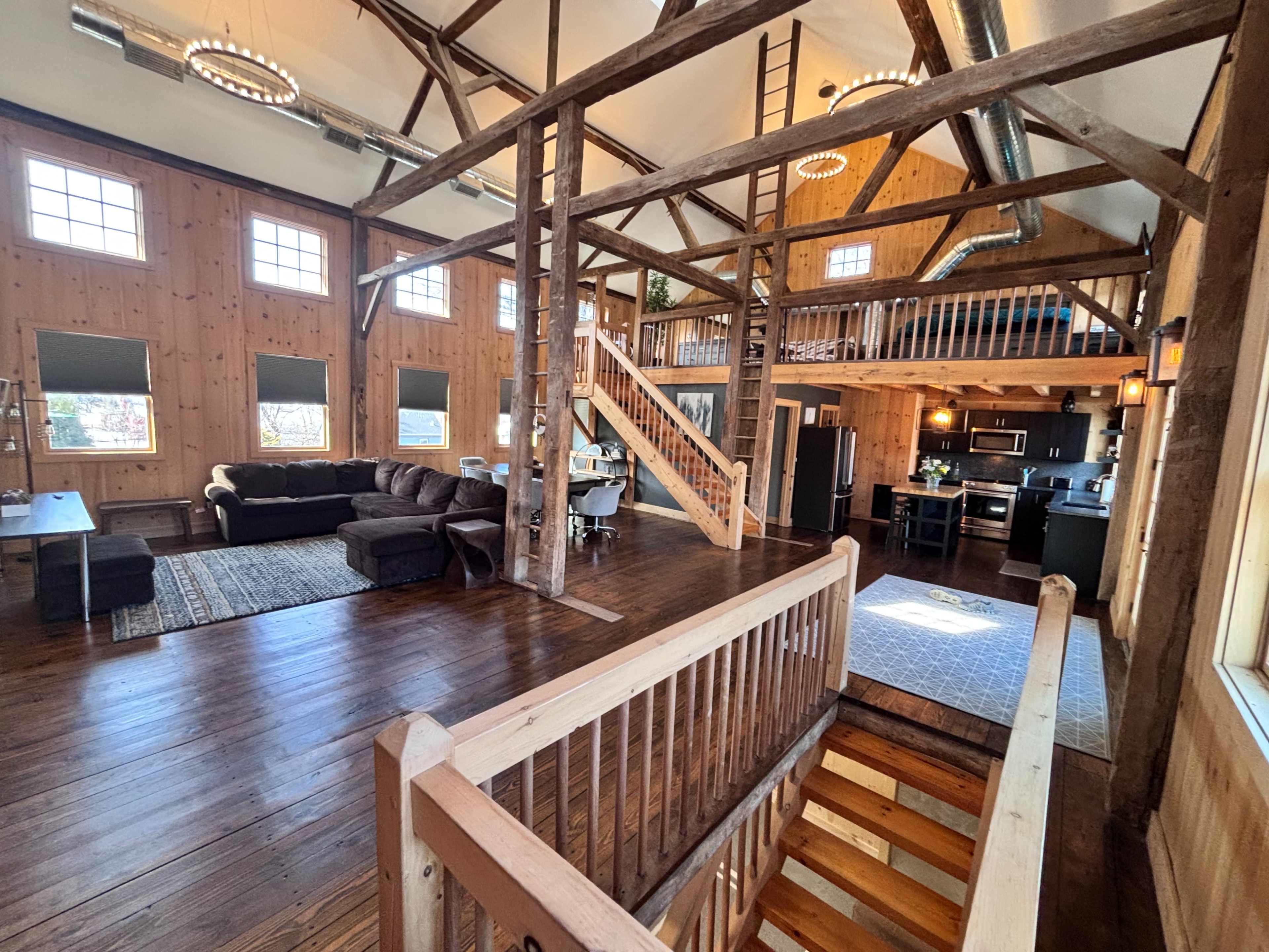 The image shows a spacious, rustic interior of a wooden cabin with open spaces, a staircase, and large windows.
